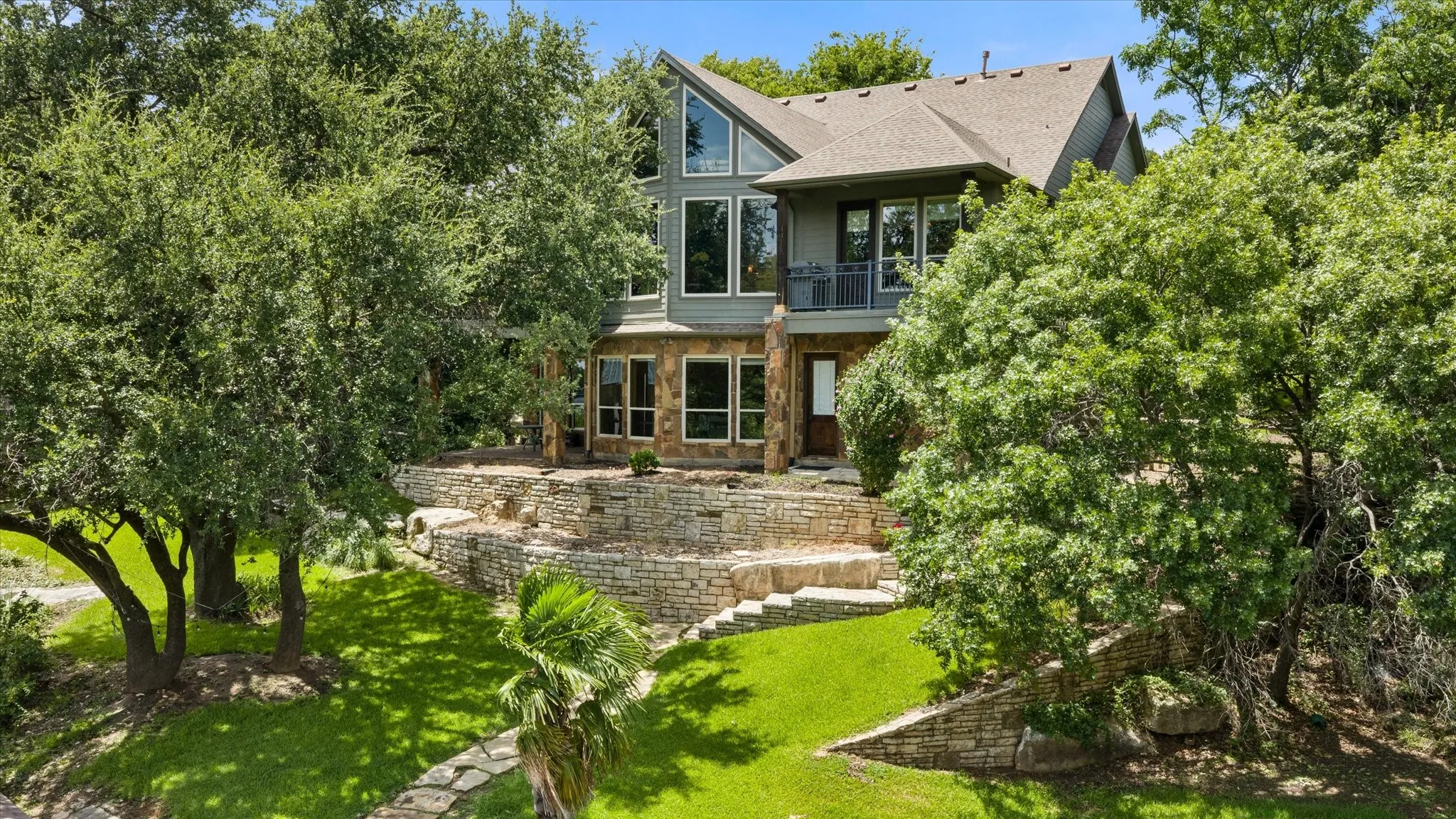 Contemporary architecture with large windows, stone facade accents, a second-story balcony, and a multi-level stone retaining wall