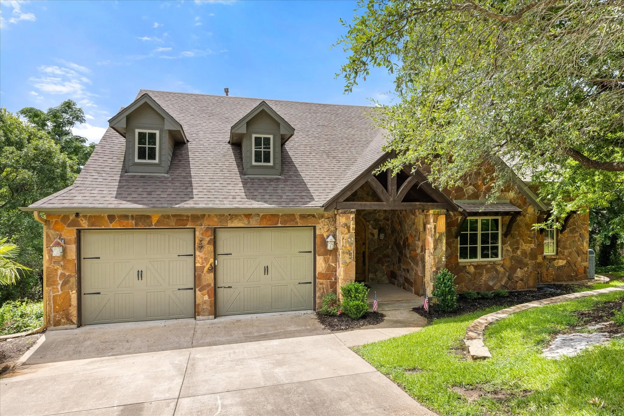 Stone-clad exterior featuring a two-car garage with carriage-style doors, a gabled porch entry with timber accents, and two dormer windows