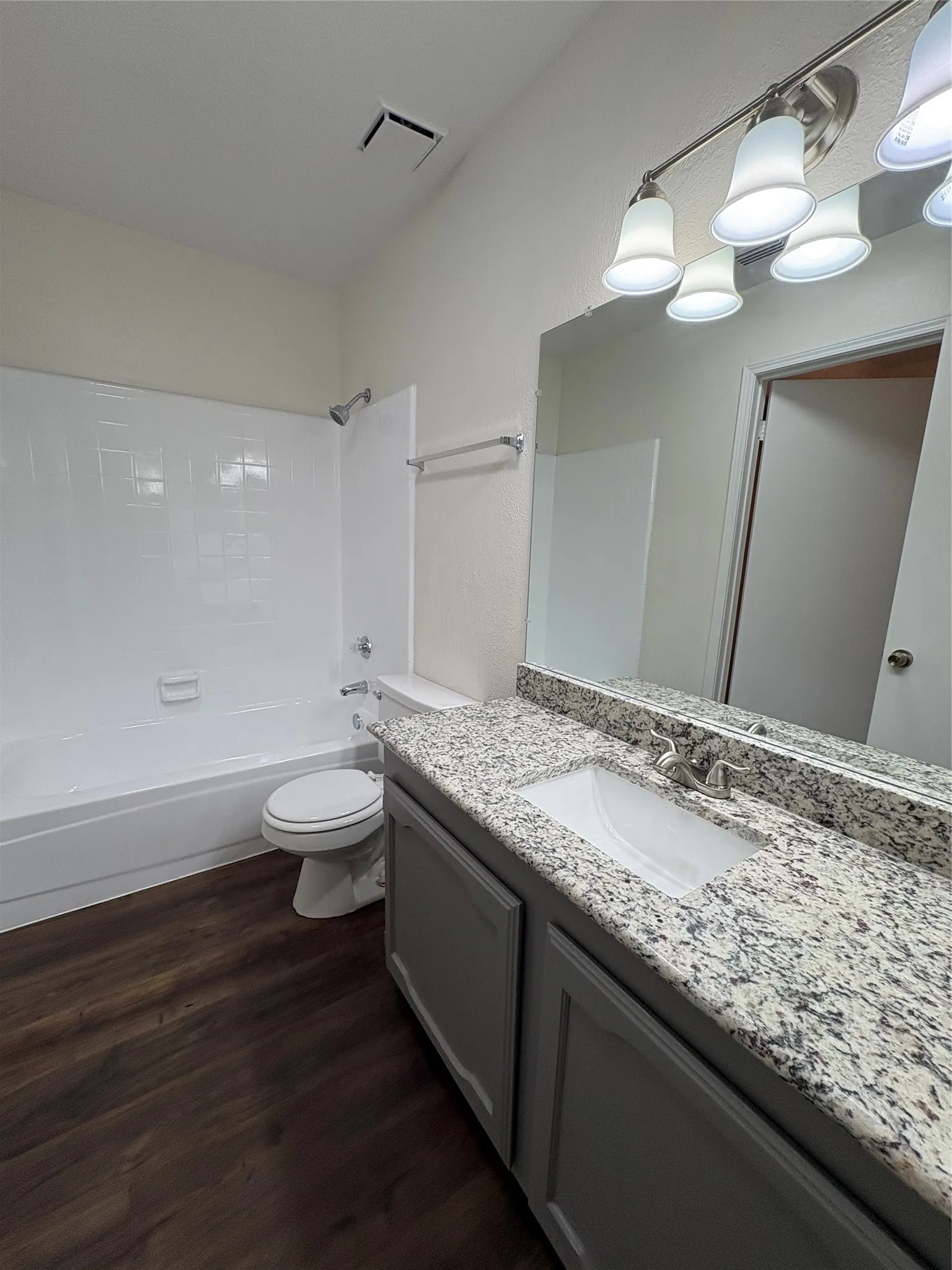 Bathroom featuring a white tub/shower combination with subway tile surround, dark wood-finish flooring, a vanity with gray cabinetry, a speckled countertop, and an undermount sink