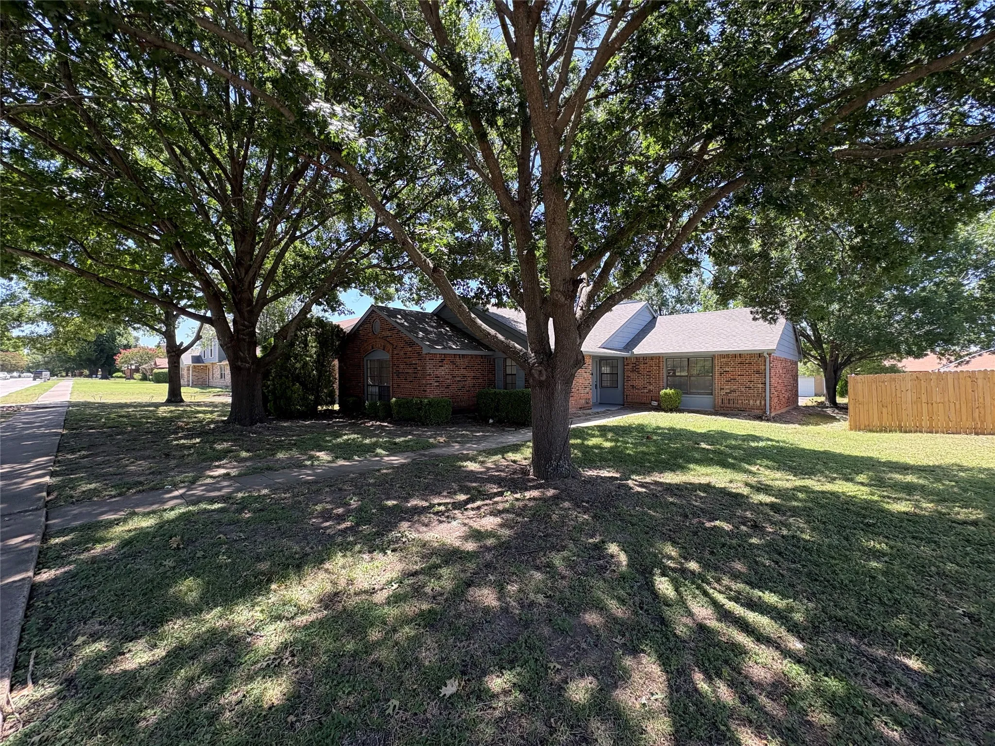 Brick exterior home featuring a gray shingle roof and a mature tree-lined front yard