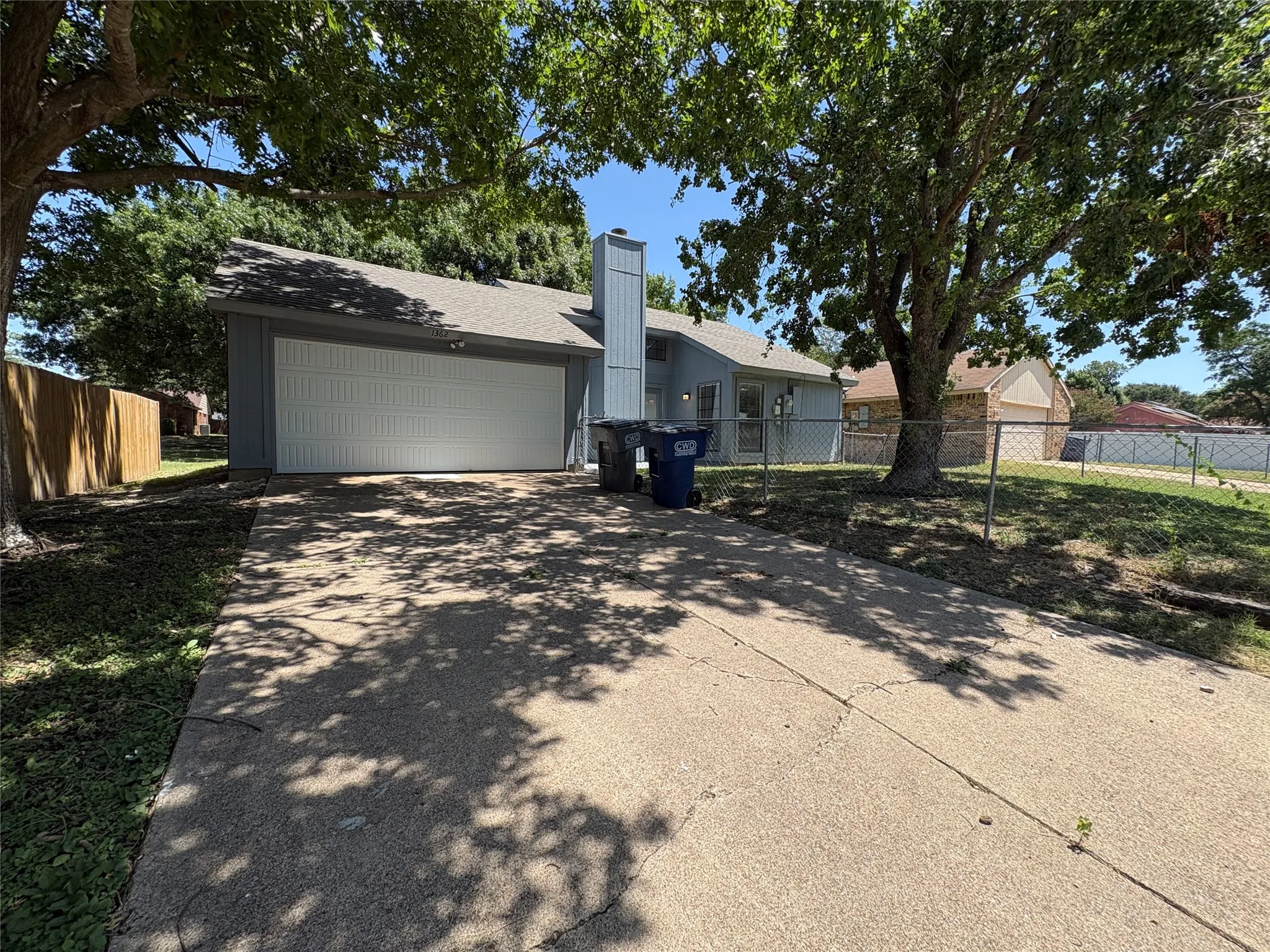 Single-story residence featuring a two-car garage with a panel door, a concrete driveway, and mature trees providing shade