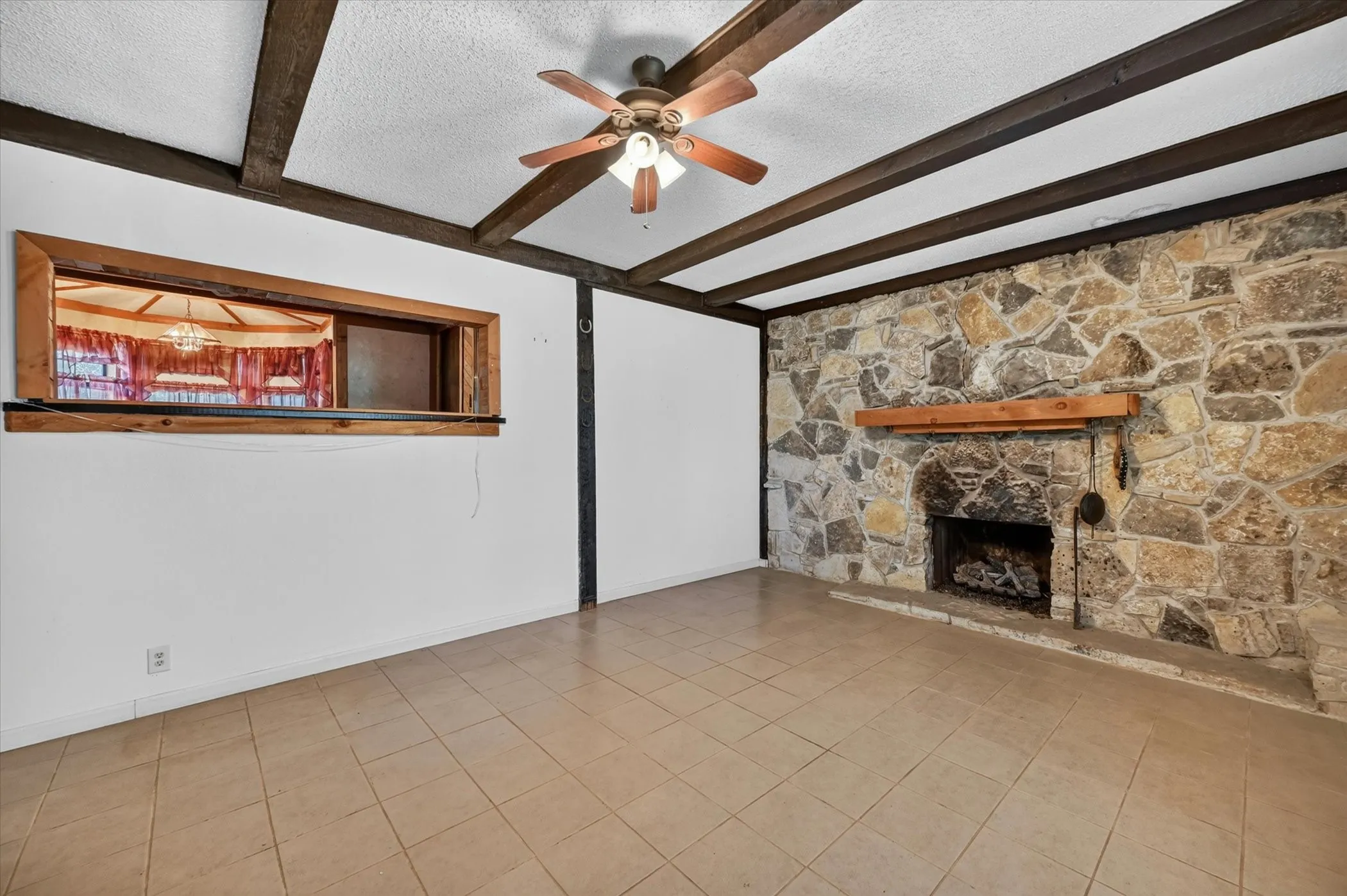 Living area featuring exposed wood beams, a stone-clad fireplace with a wood mantle, tile flooring, and a pass-through window