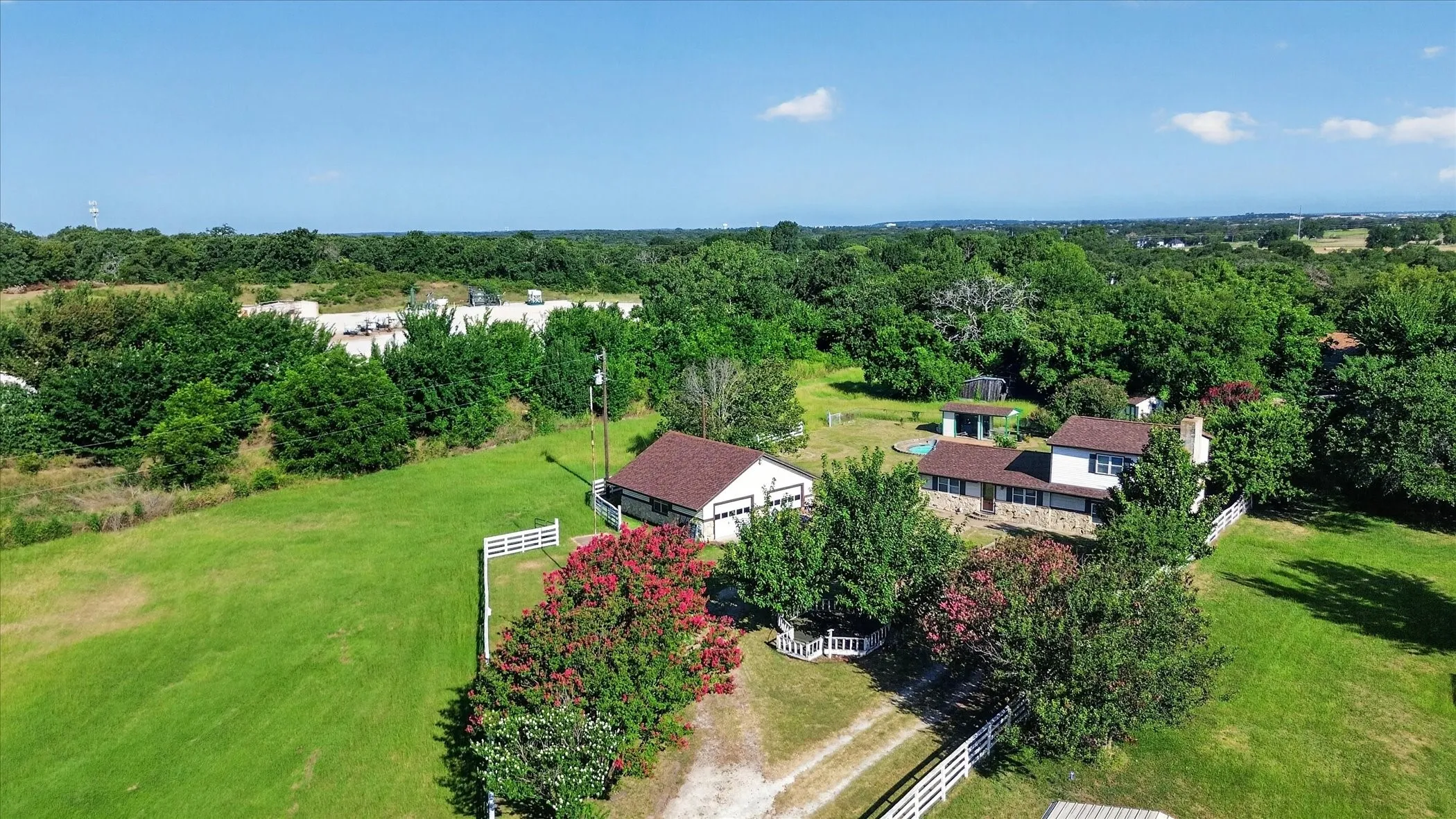 Expansive property featuring a two-story residence with a stone facade, a separate outbuilding with a brown shingle roof, lush green lawns, mature trees, and a white fence bordering the property