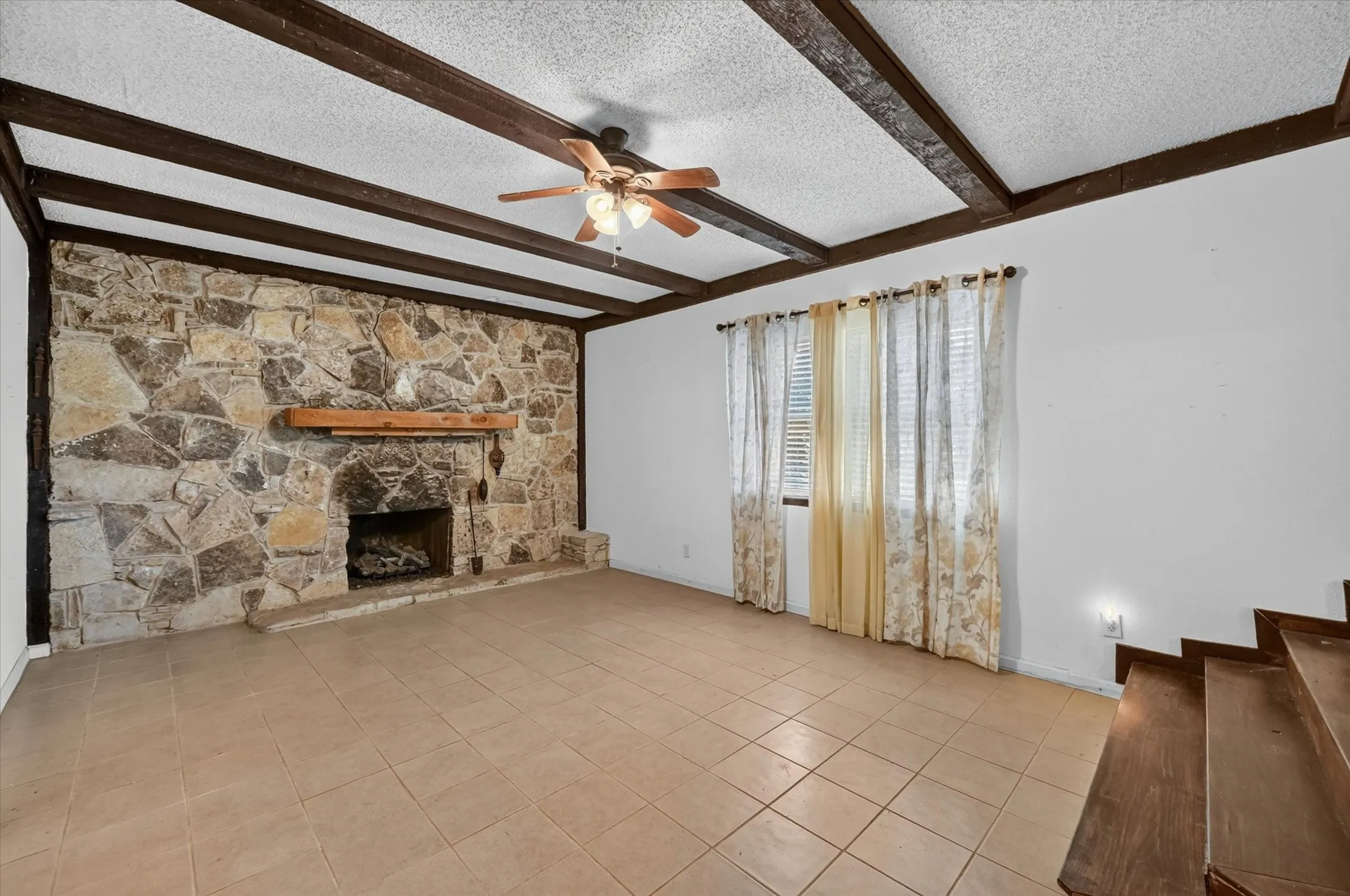 Spacious room featuring a stone-faced fireplace with a wood mantel, exposed ceiling beams, a ceiling fan with integrated lighting, and tile flooring