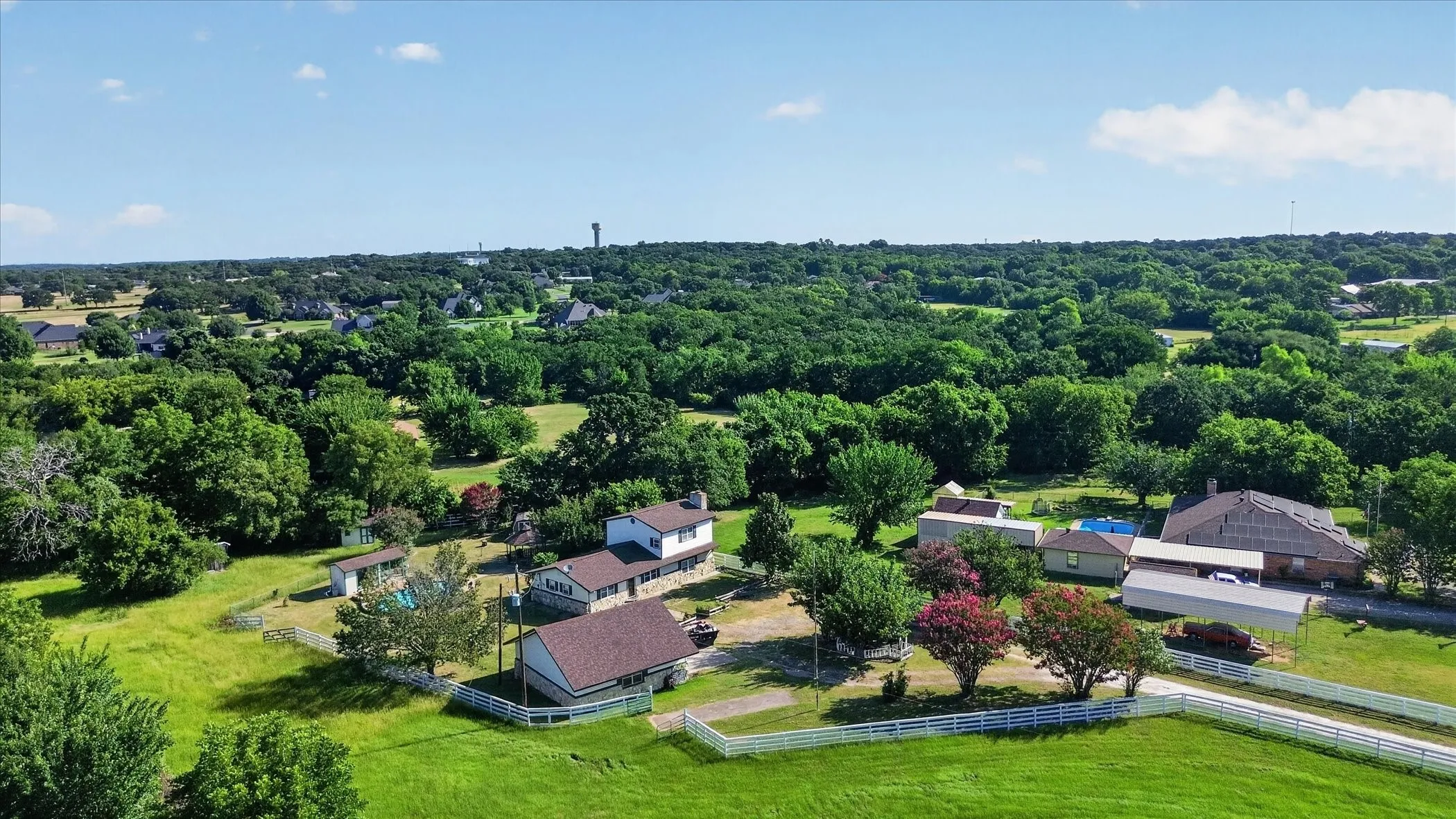 Expansive property featuring multiple structures, mature trees, a white picket fence, and a distant water tower