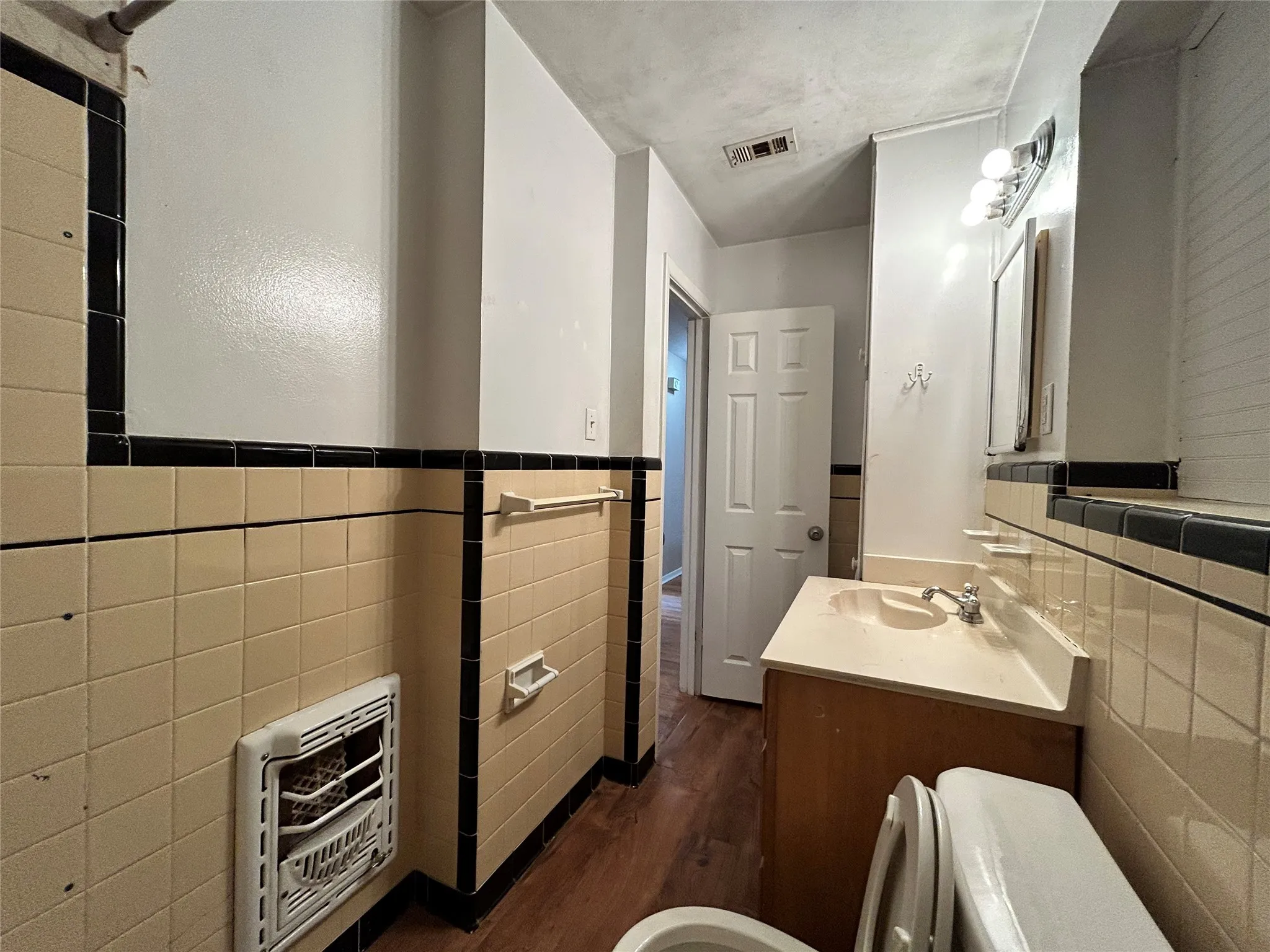 Wood-finish flooring, tiled walls with dark accent trim, vanity with integrated sink, wall-mounted light fixture, and a white paneled door