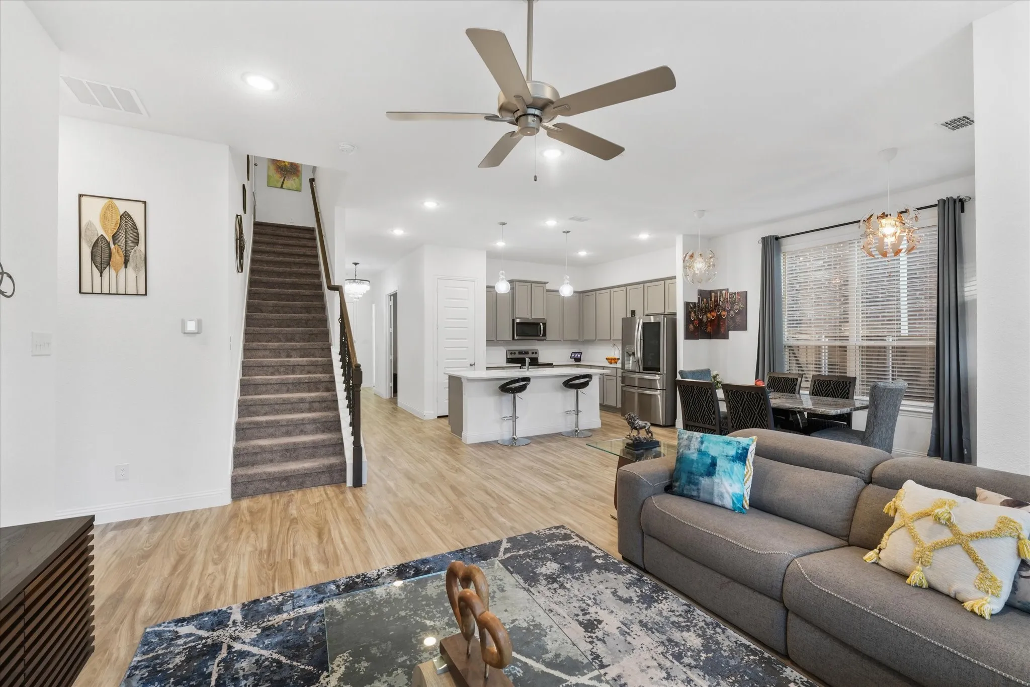 Open-concept living space featuring wood-finish flooring, a contemporary kitchen with stainless steel appliances, a breakfast bar island with seating, a dining area, and a carpeted staircase with dark wood railings