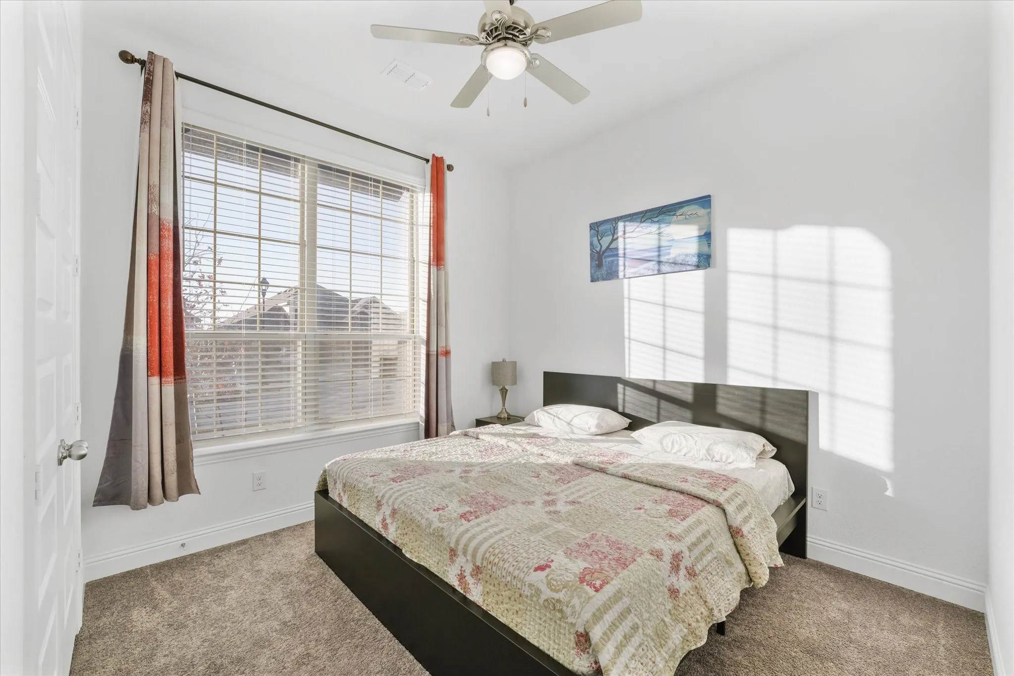 Bright bedroom featuring a large window with blinds, a ceiling fan with integrated lighting, textured carpeting, and white baseboards