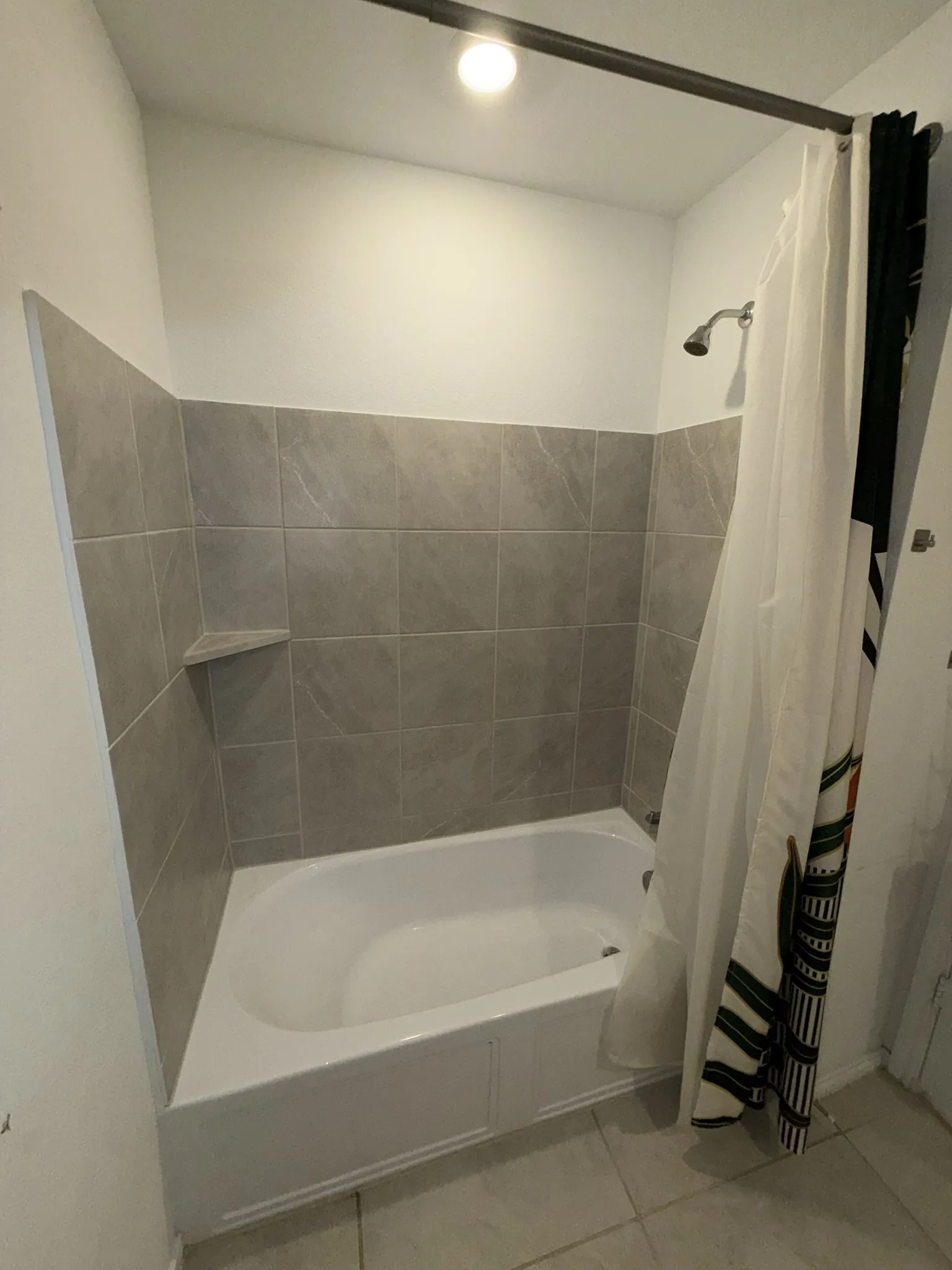 Bathtub and shower combination featuring gray tile surround, integrated corner shelf, and a ceiling-mounted light fixture