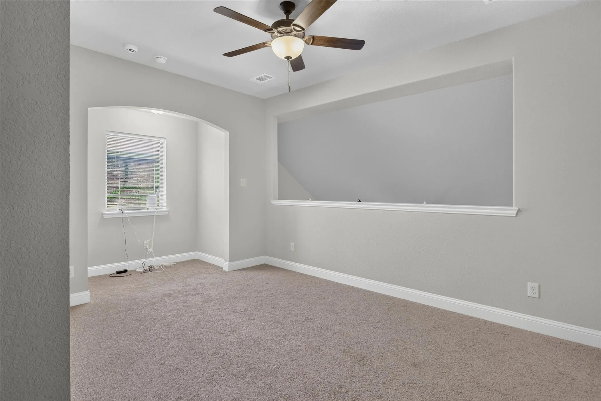 Spacious room featuring neutral carpet, light gray walls, a window with horizontal blinds, an arched wall opening, and a ceiling fan with integrated lighting