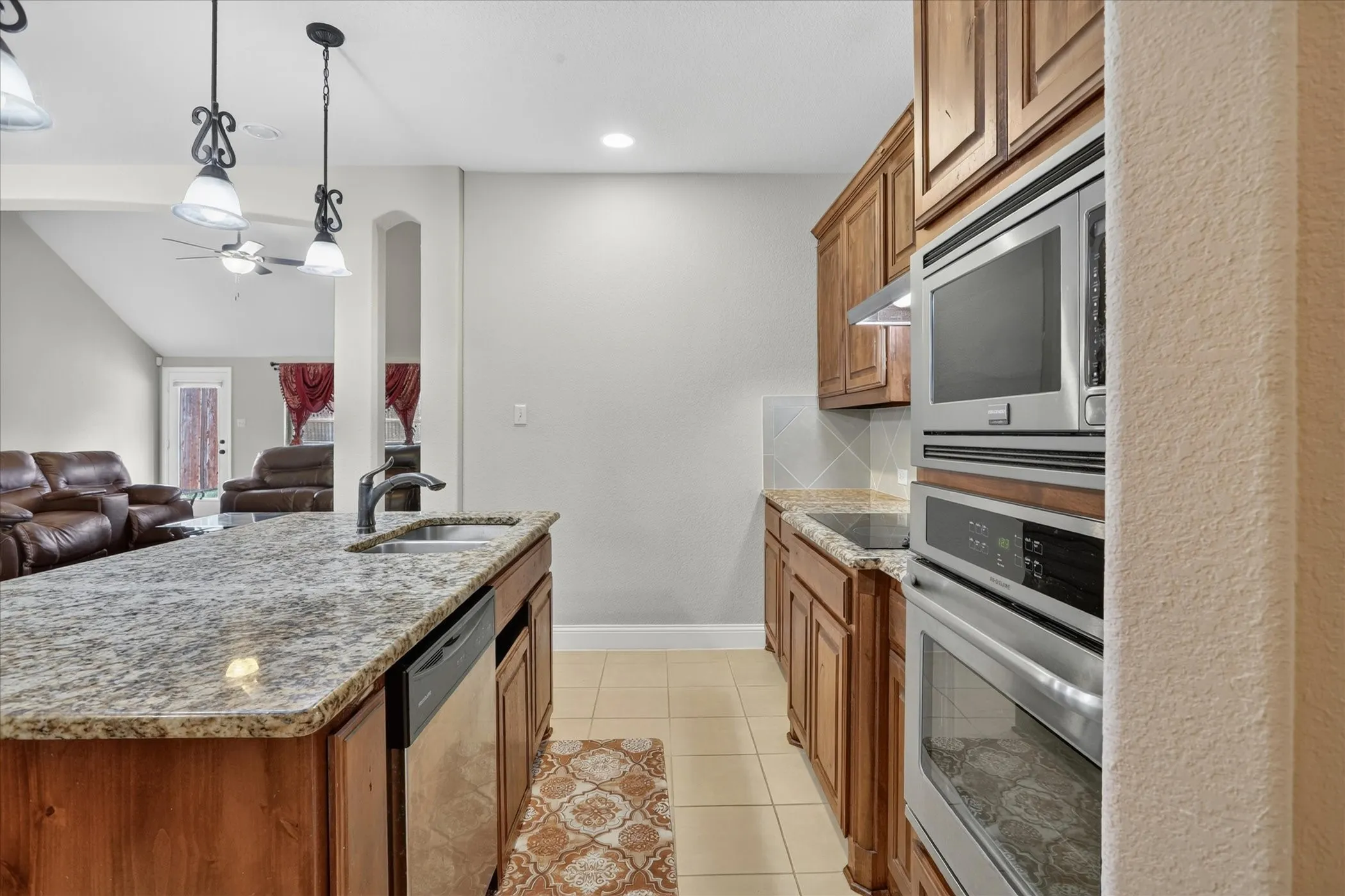 Kitchen featuring a granite island with an undermount sink, pendant lighting, built-in stainless steel appliances, wood cabinetry, and tile flooring