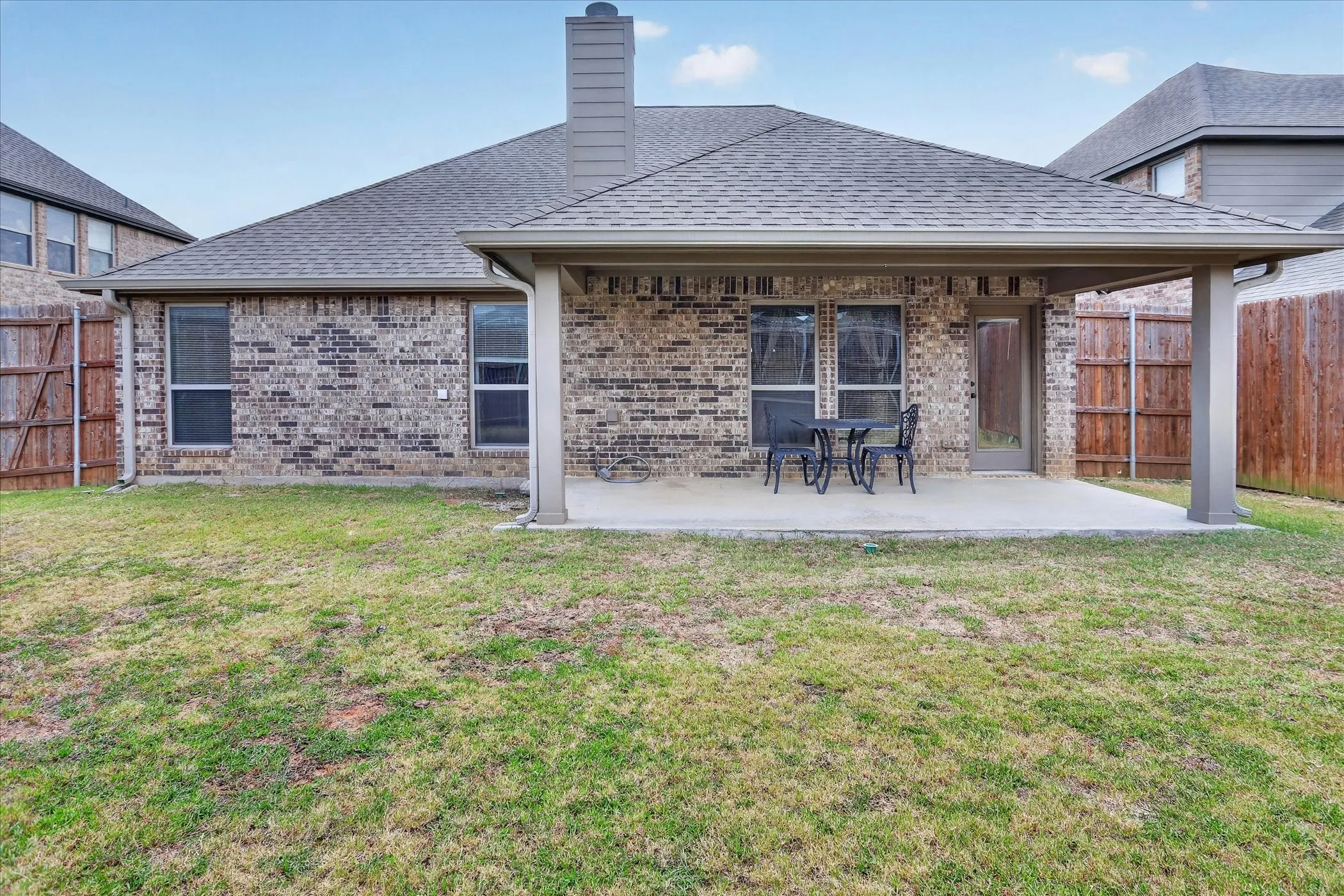 Expansive grass yard with a brick exterior, covered patio, multiple windows, and a chimney