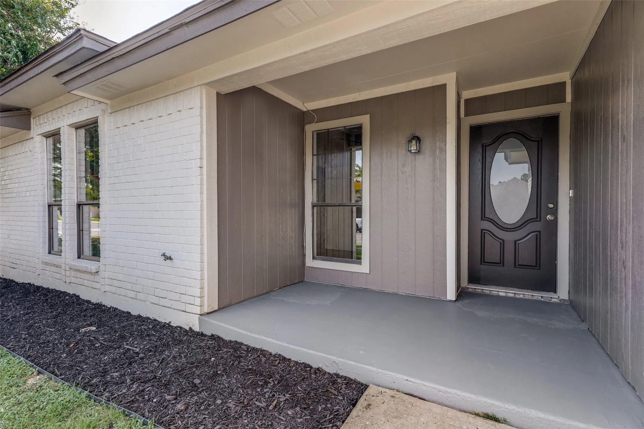 Covered front porch featuring a dark wood-finish door with an oval glass insert, a complementary wall-mounted light fixture, and vertical siding