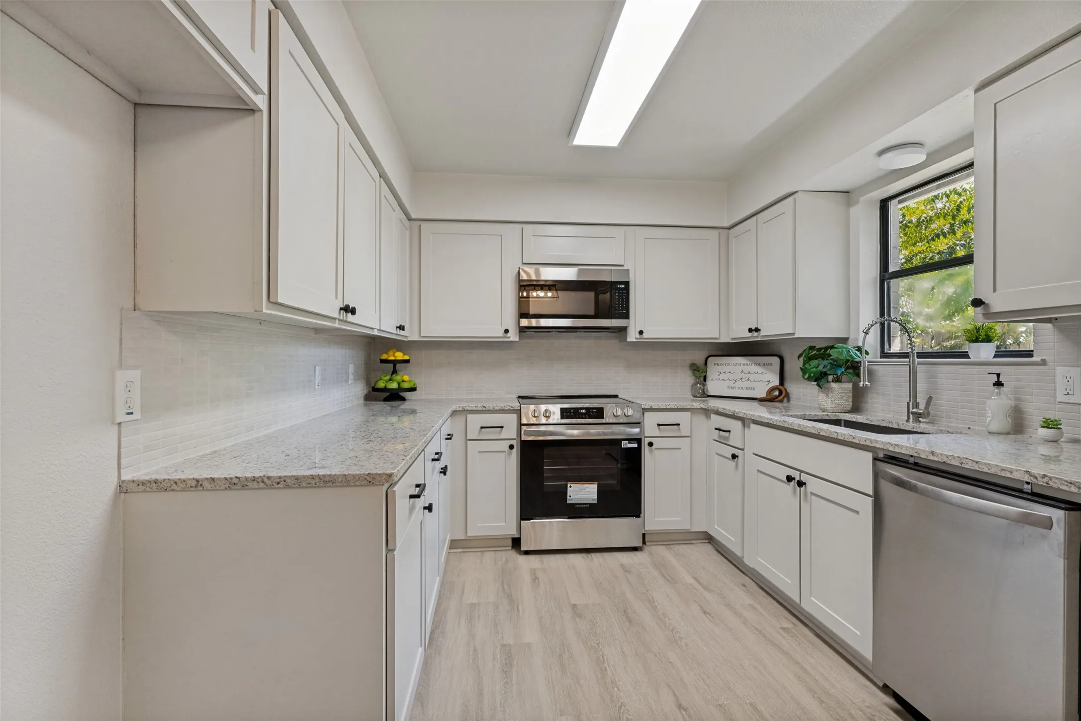U-shaped kitchen with white shaker-style cabinetry, light wood-finish flooring, granite-style countertops, stainless steel appliances, and a mosaic tile backsplash
