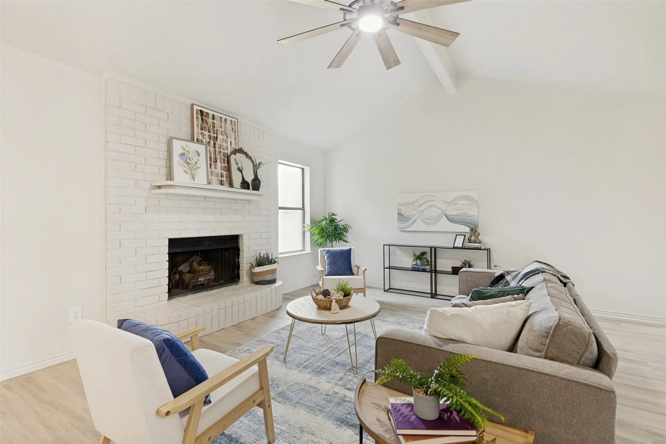 Living area featuring a painted brick fireplace with a mantel, vaulted ceiling, wood-finish flooring, and a ceiling fan with integrated lighting