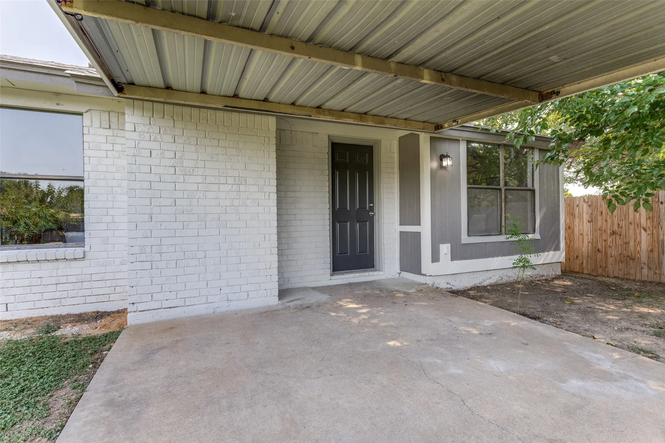 Carport entry featuring a corrugated metal roof, painted brick facade, dark paneled entry door, and exterior wall lantern