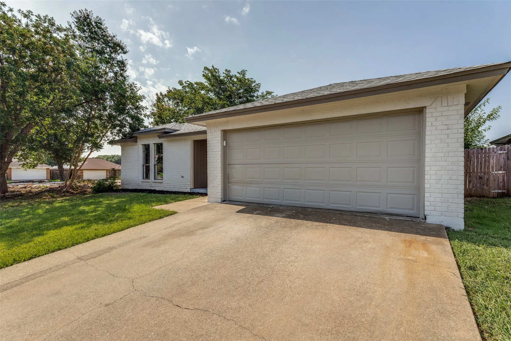 Exterior featuring white painted brick, a two-car garage with a panel door, a concrete driveway, and mature trees