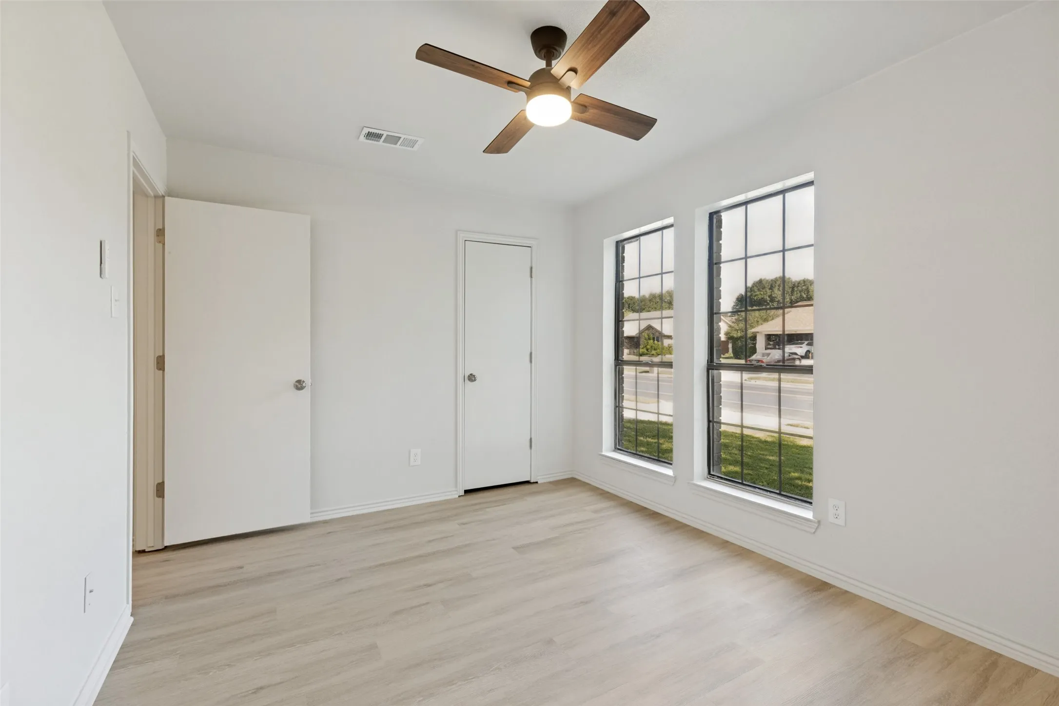 Spacious room featuring light wood-finish flooring, a ceiling fan with light fixture, two tall windows with grid patterns, and two white interior doors