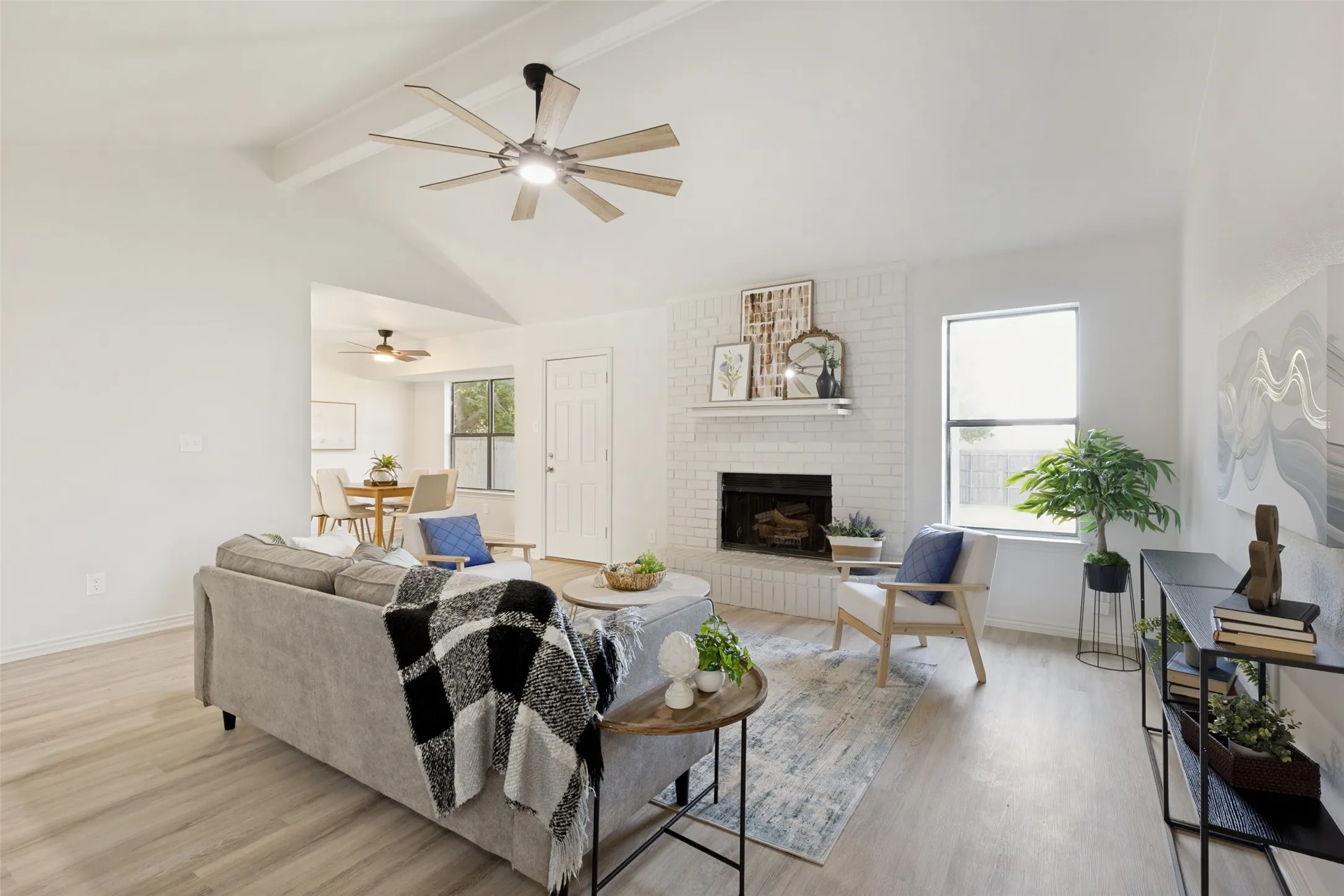 Vaulted ceiling living area featuring light wood-finish flooring, a white-painted brick fireplace with mantle, and a contemporary ceiling fan