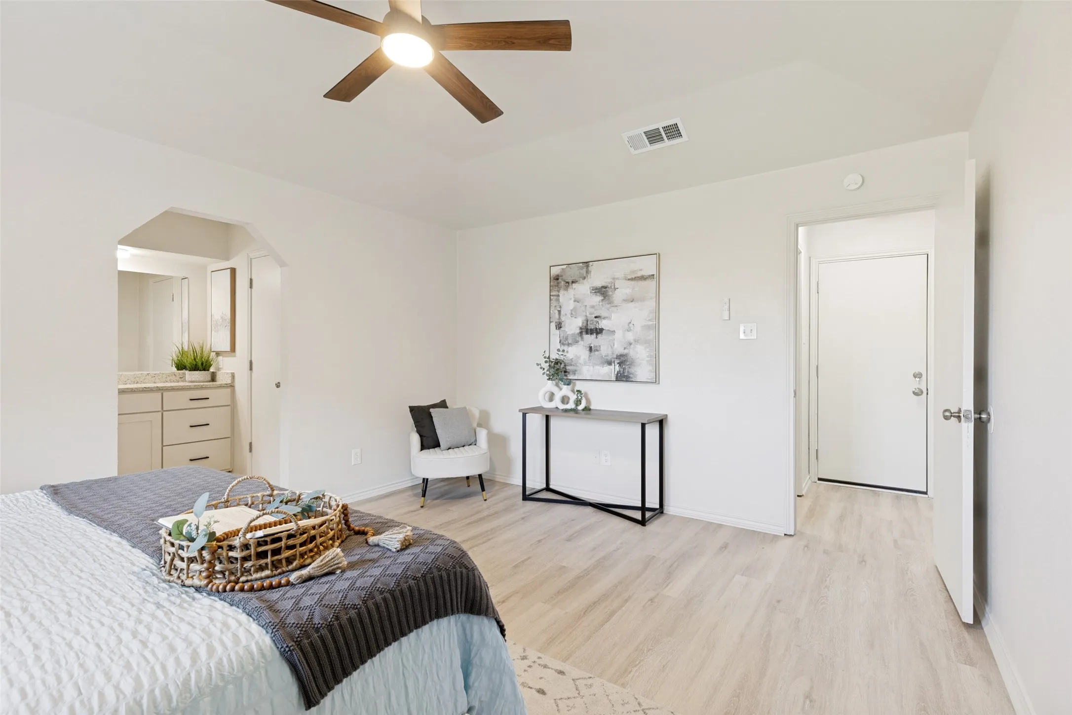 Spacious room with light wood-finish flooring, a ceiling fan, and an arched entry leading to a vanity area with a light-toned cabinet and countertop