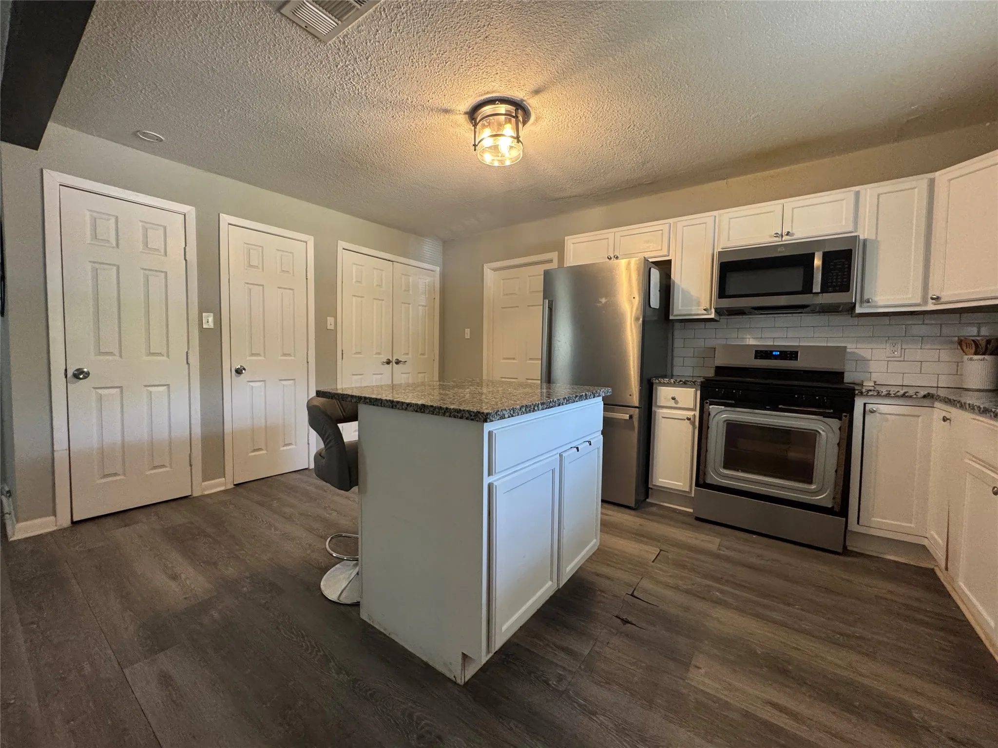 Kitchen featuring a central island with a granite-style countertop, white cabinetry, wood-finish flooring, stainless steel appliances, and a white subway tile backsplash