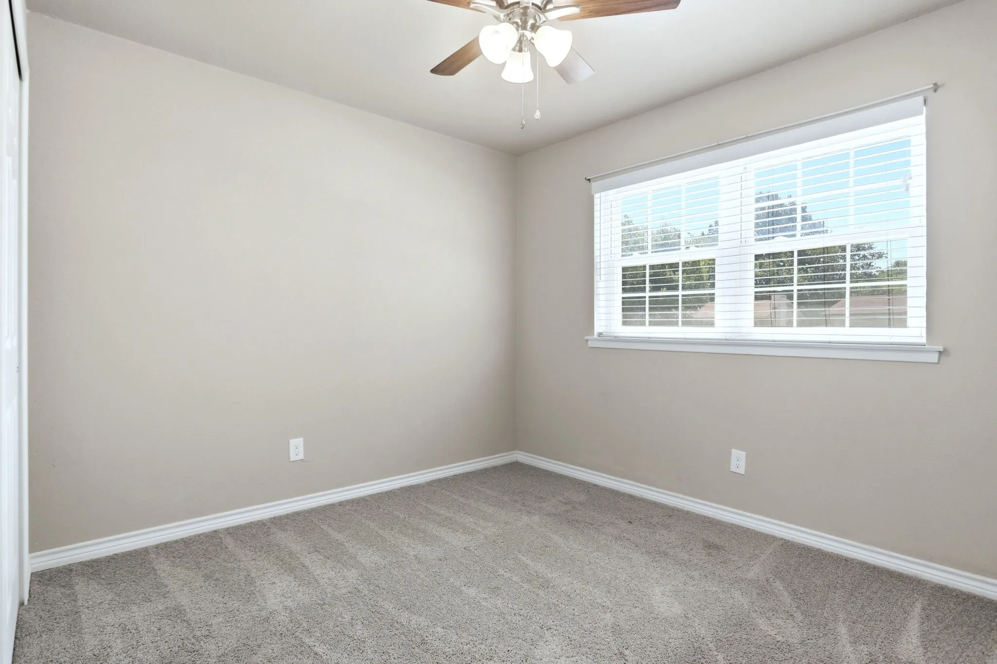 Carpeted room featuring a double window with blinds, a ceiling fan with wood-finish blades, light-colored walls, and white baseboards