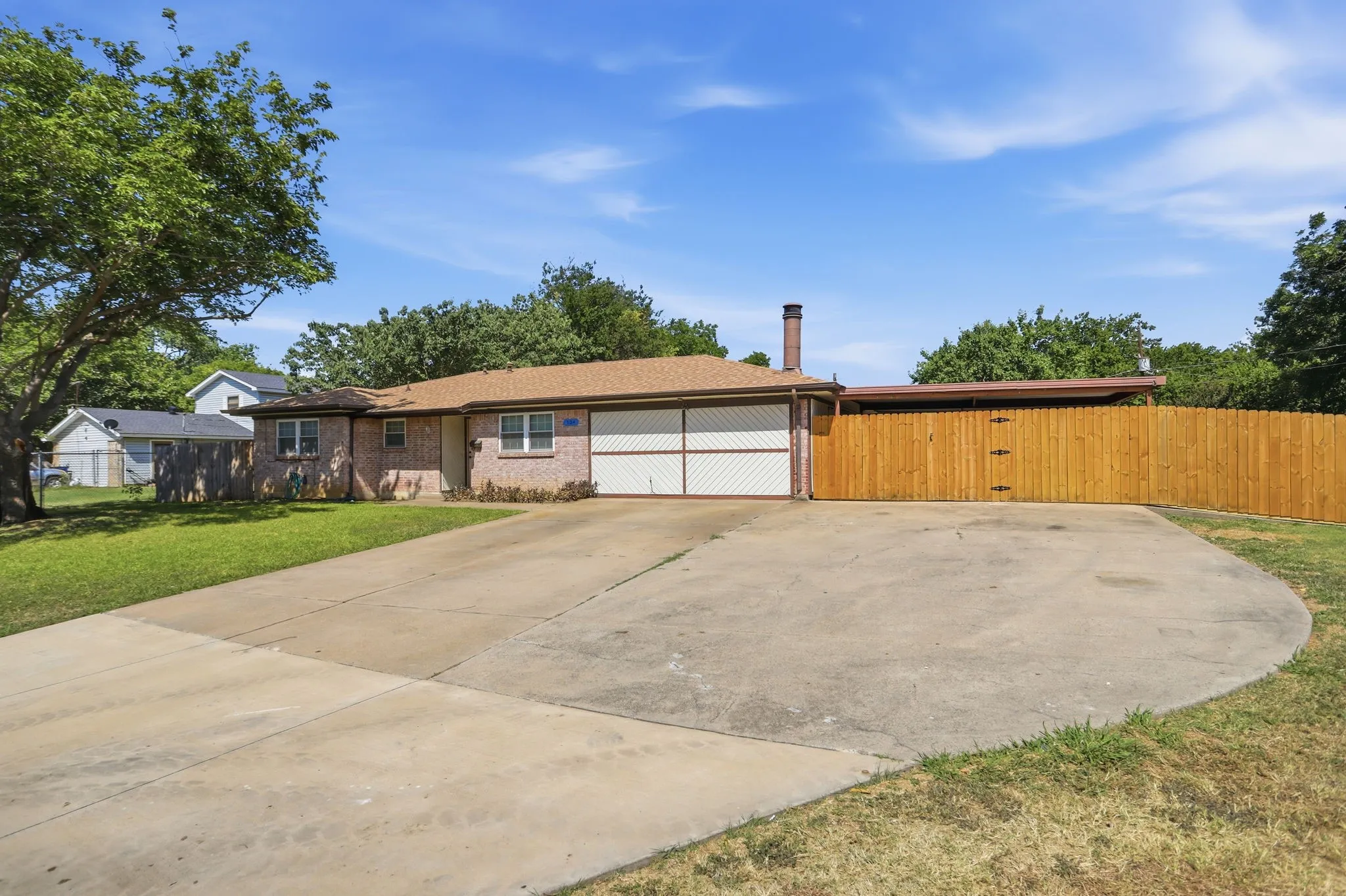 Brick facade residence featuring an attached two-car garage, a wide concrete driveway, a new wooden privacy fence, and mature trees
