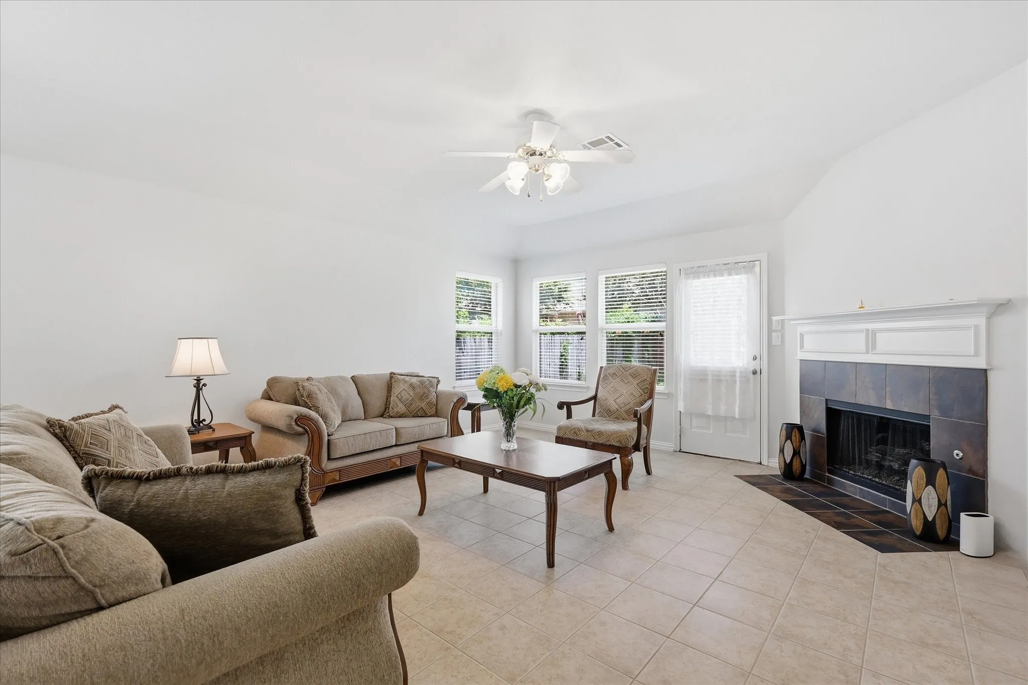 Spacious living area featuring neutral-toned tile flooring, a vaulted ceiling, and a fireplace with a white mantel and dark tile surround