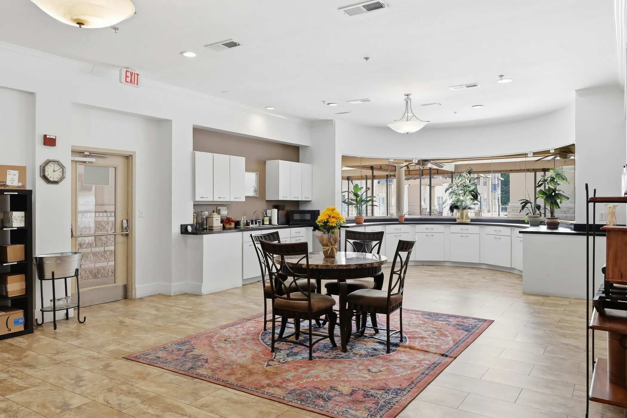 Spacious open common area featuring light-toned tile flooring, recessed lighting, a built-in kitchenette with white cabinetry and dark countertops, and a prominent curved wall of large windows