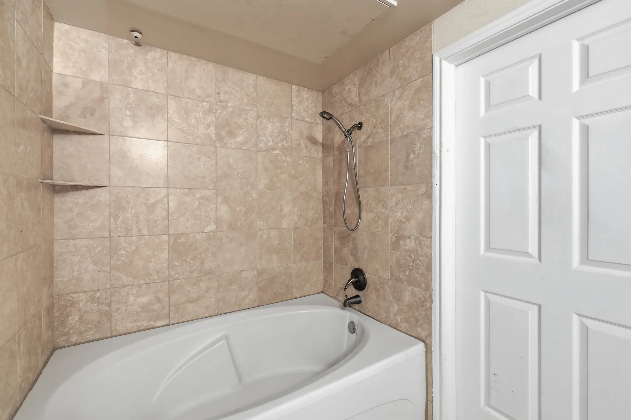 Alcove bathtub featuring a handheld showerhead, dark bronze fixtures, and natural-tone tile surround