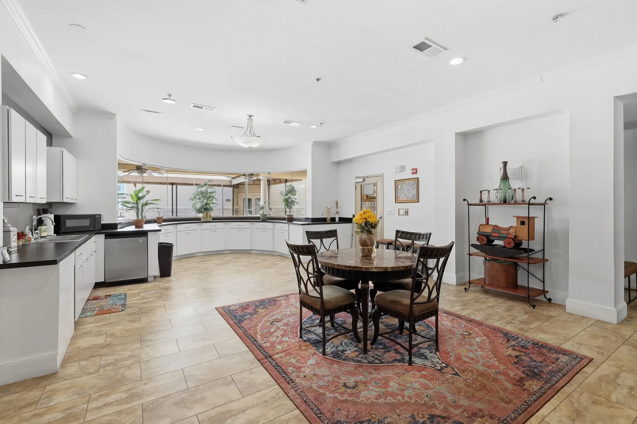 Spacious common area featuring extensive white cabinetry, dark countertops, tile flooring, a dining table with chairs, and a curved window wall