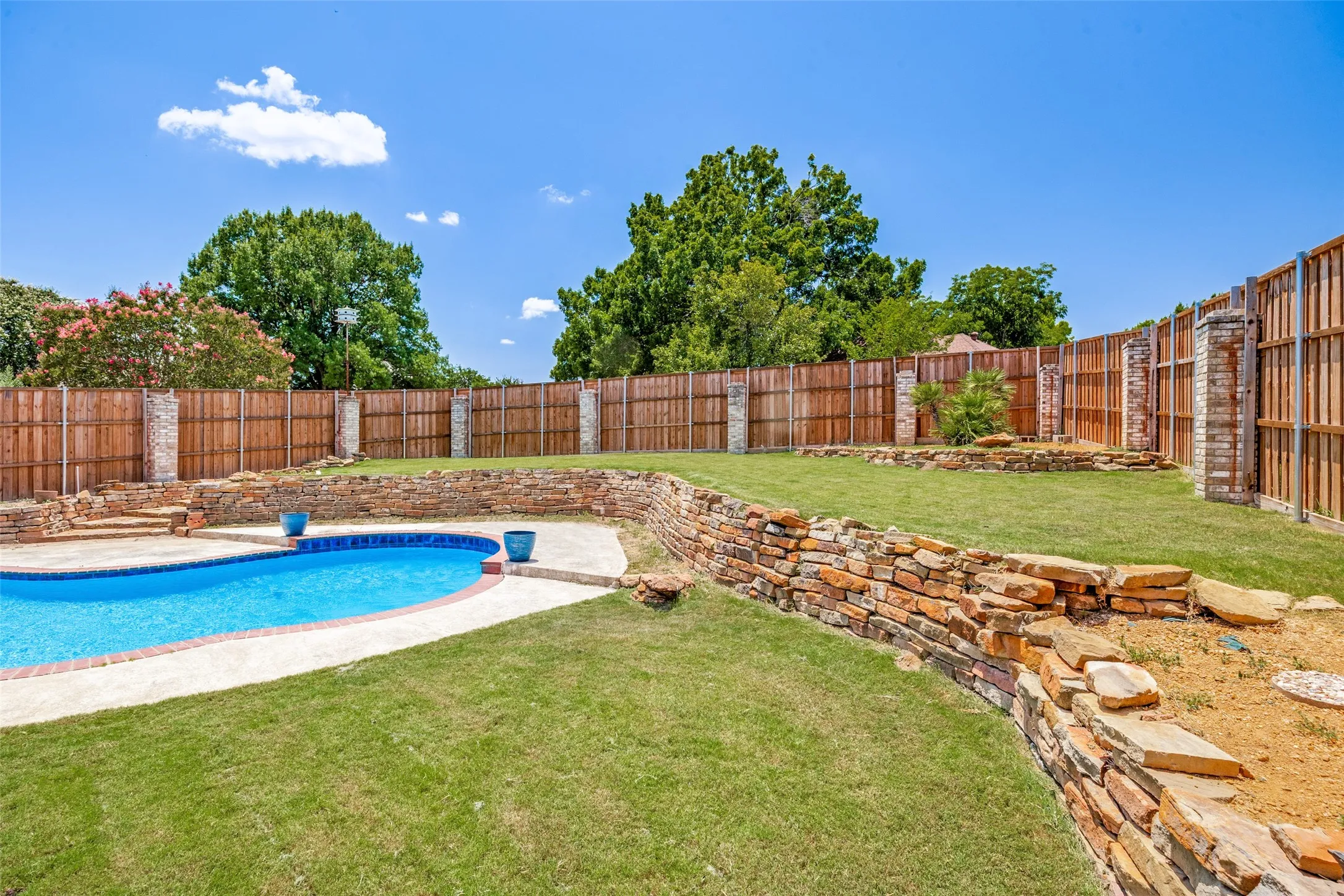 Curved inground pool with concrete decking, surrounded by a tiered backyard featuring stone retaining walls, green lawn, and a wood privacy fence with brick pillars