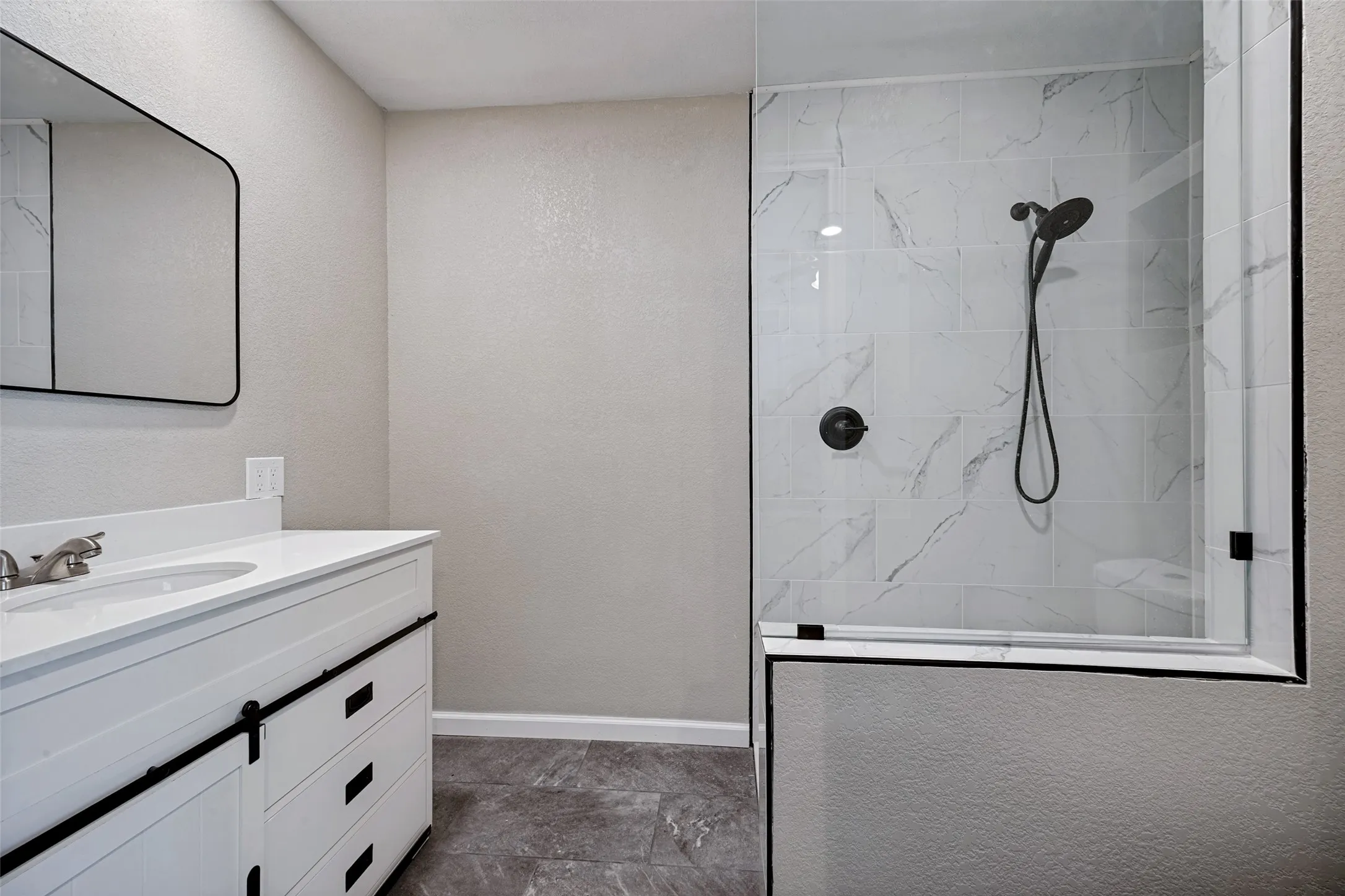 Contemporary bathroom featuring a frameless glass shower enclosure with white marble-look tile, a matte black shower system, a white vanity with barn door style hardware, and a coordinating rectangular mirror