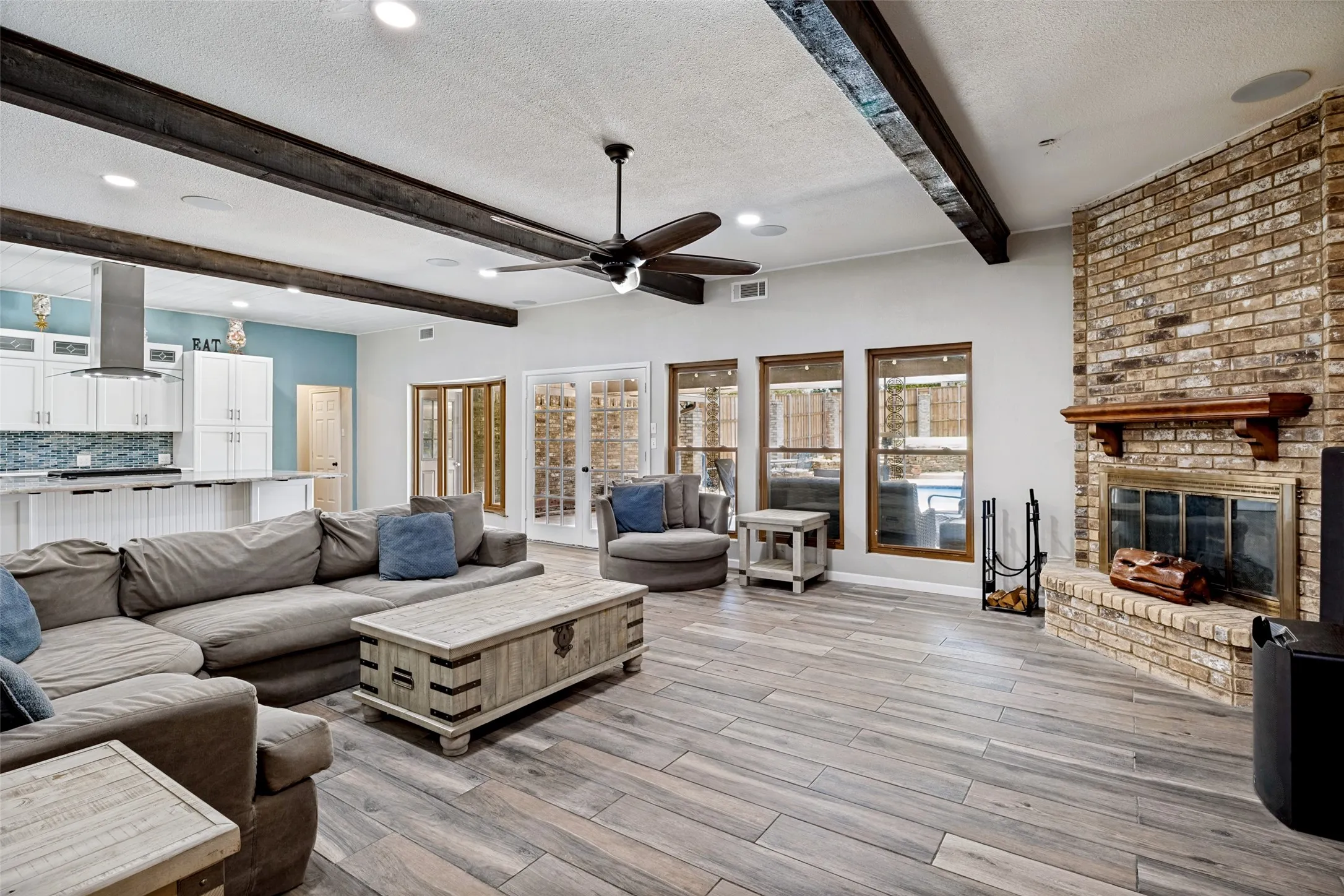 Open concept living space featuring porcelain ceramic wood-finish flooring, exposed ceiling beams, and a brick fireplace