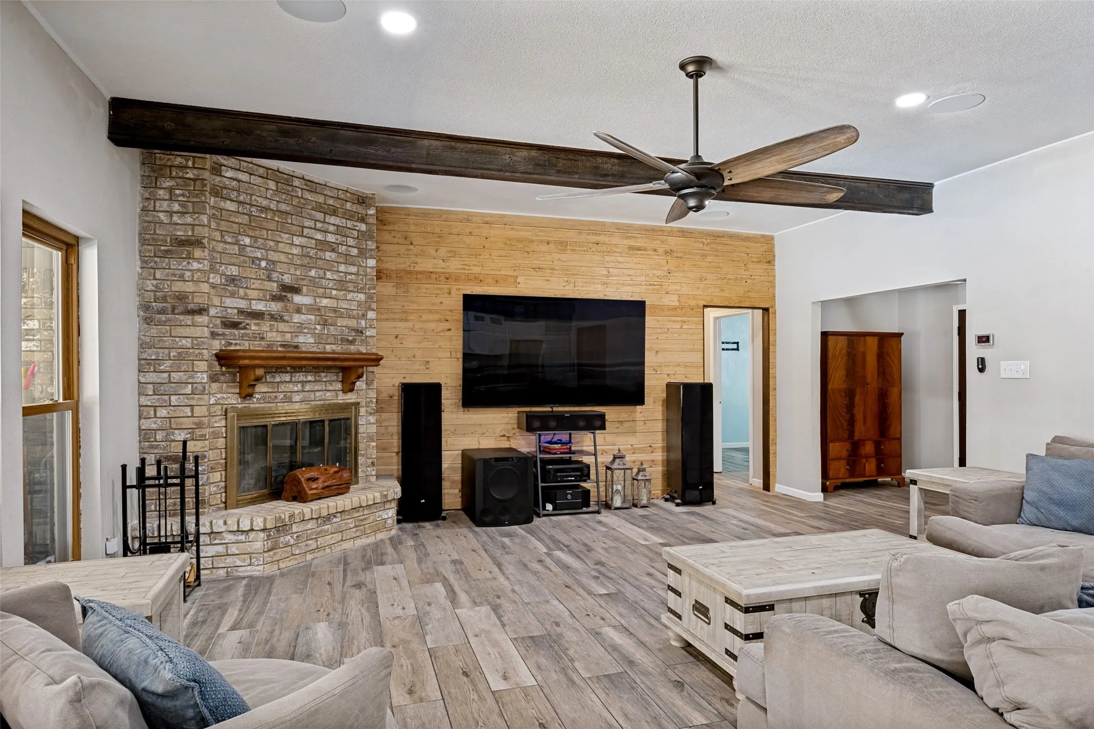 Spacious living area featuring porcelain ceramic wood-finish flooring and a corner brick fireplace with a wood​​‌​​​​‌​​‌‌​‌‌​​​‌‌​‌​‌​‌​​​‌​​ mantle