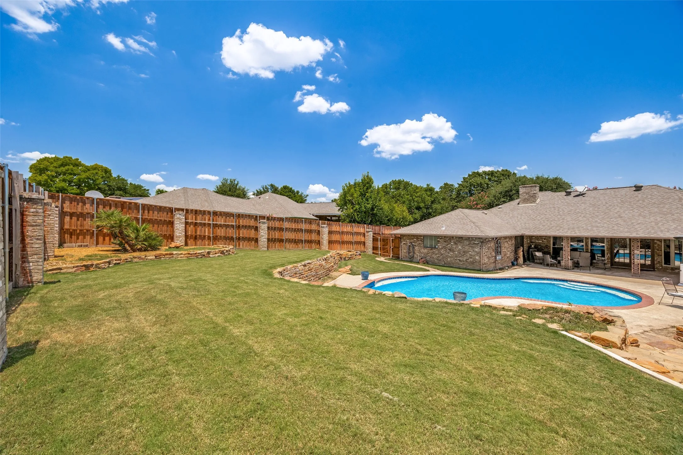 Expansive rear yard featuring a freeform swimming pool, stone patio, and a privacy fence with lighted brick columns