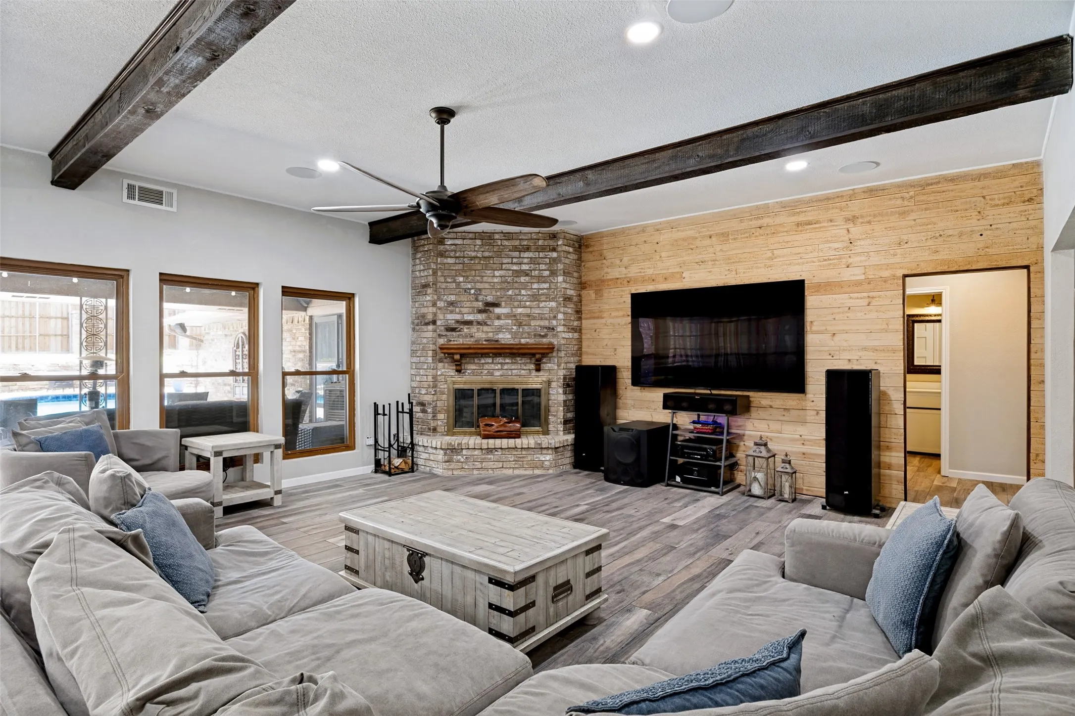 Spacious living area featuring exposed wood-finish ceiling beams, porcelain ceramic wood-finish flooring, and a brick-finish fireplace with a wood mantel