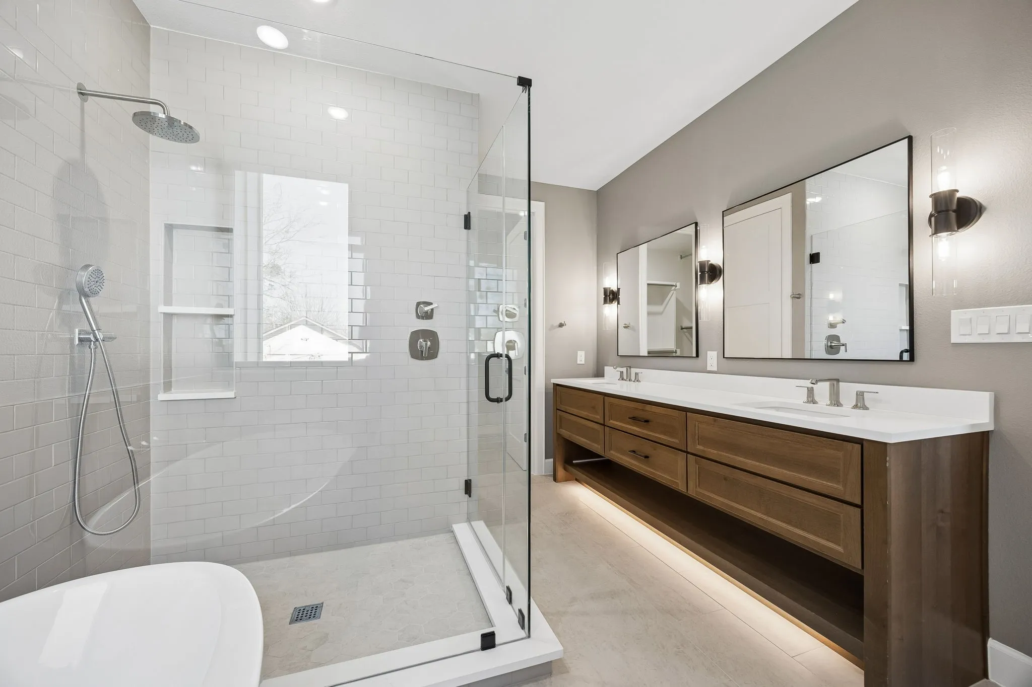 Spacious bathroom featuring a glass-enclosed shower with a rain showerhead and hand-held sprayer, dual vanity with wood-finish cabinetry and undermount sinks, and integrated lighting