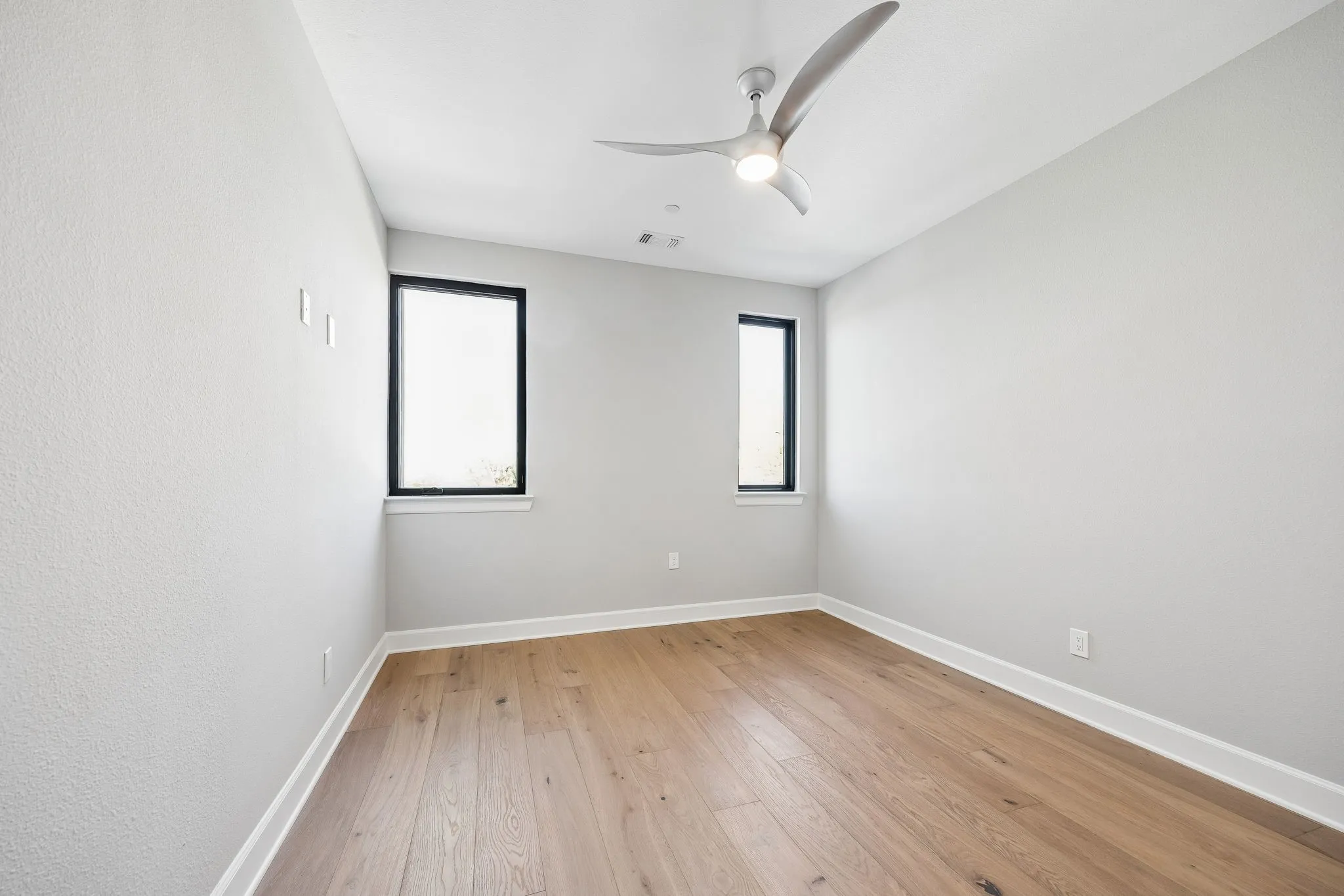 Room featuring light wood-finish flooring, two rectangular black-framed windows, and a modern ceiling fan with integrated lighting