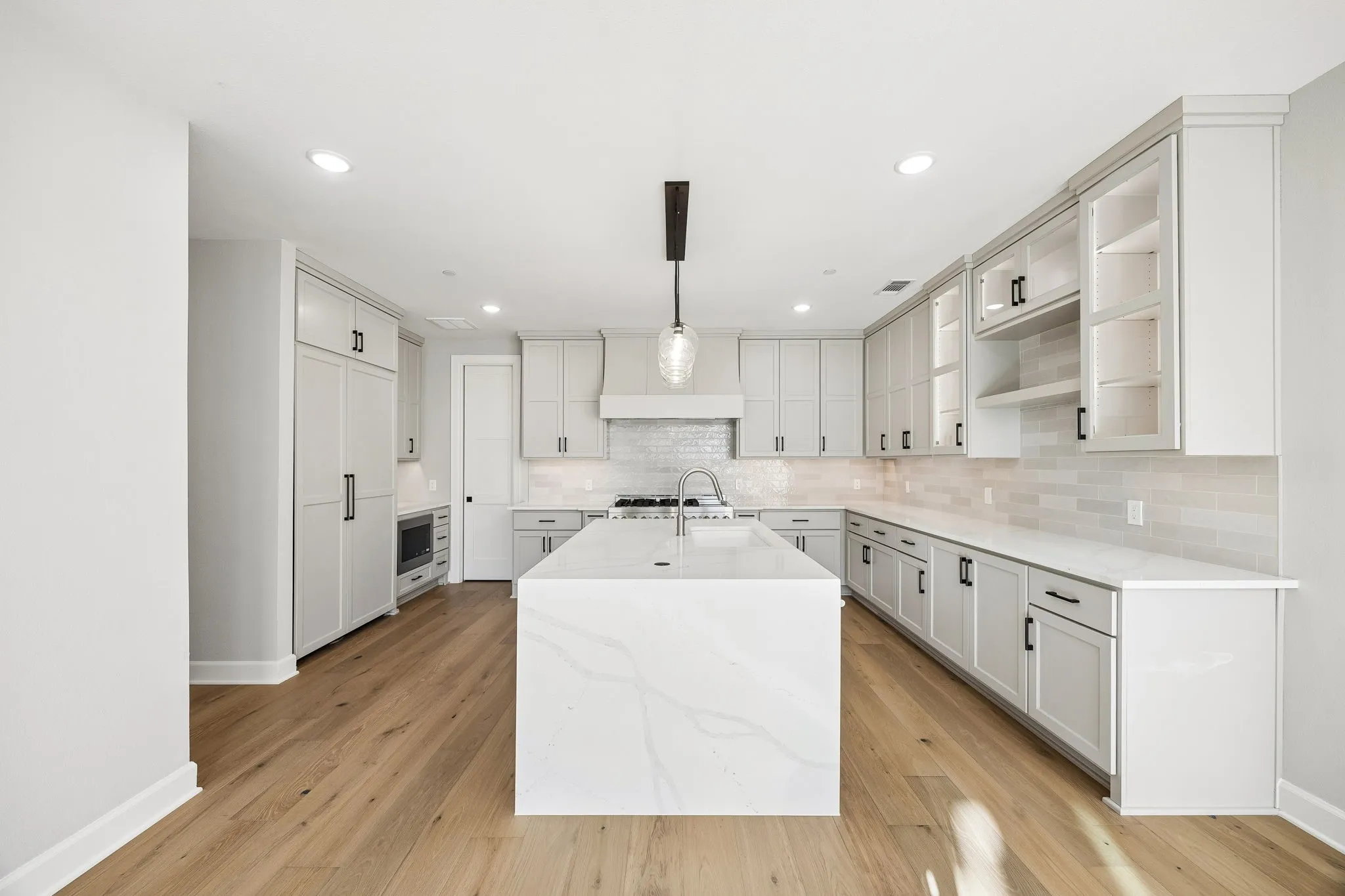 Spacious kitchen featuring a large central island, light wood-finish flooring, white shaker cabinetry with dark hardware, quartz countertops, and an integrated microwave