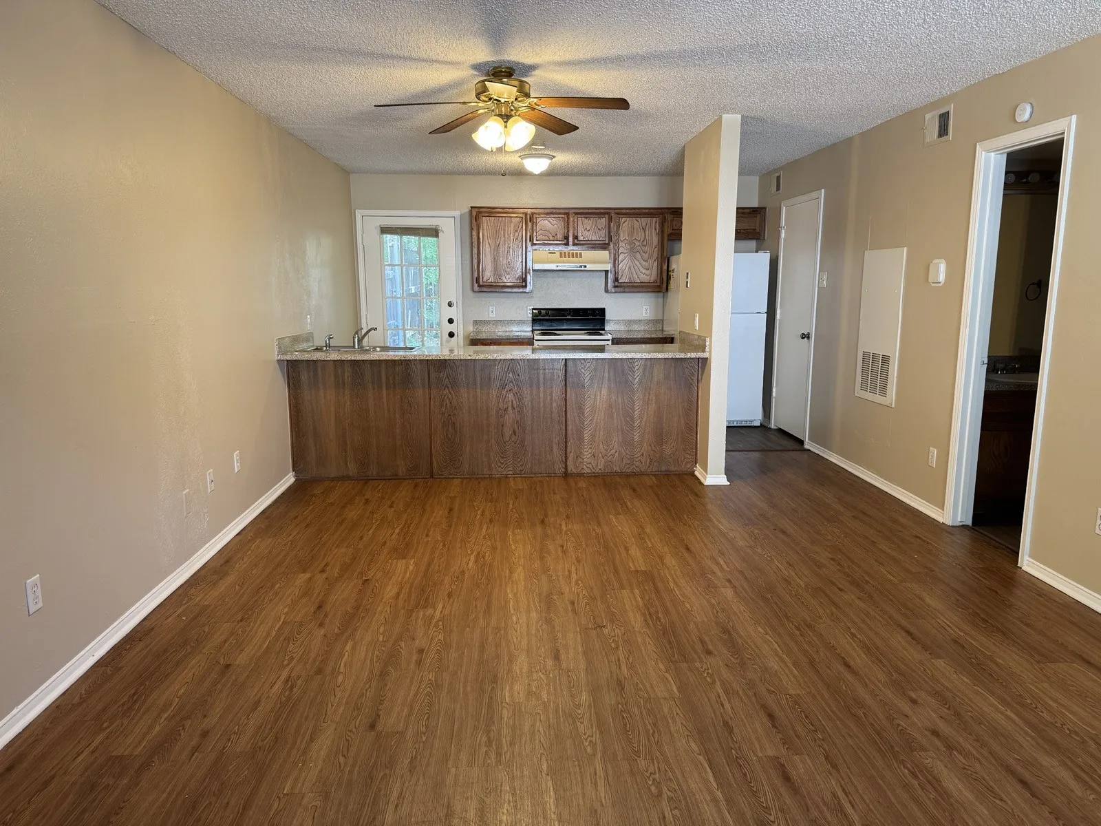 Open-concept living area featuring wood-finish flooring, a kitchen with wood cabinetry, a ceiling fan with integrated lighting, a breakfast bar with a stone-look countertop, and a glass-paned door