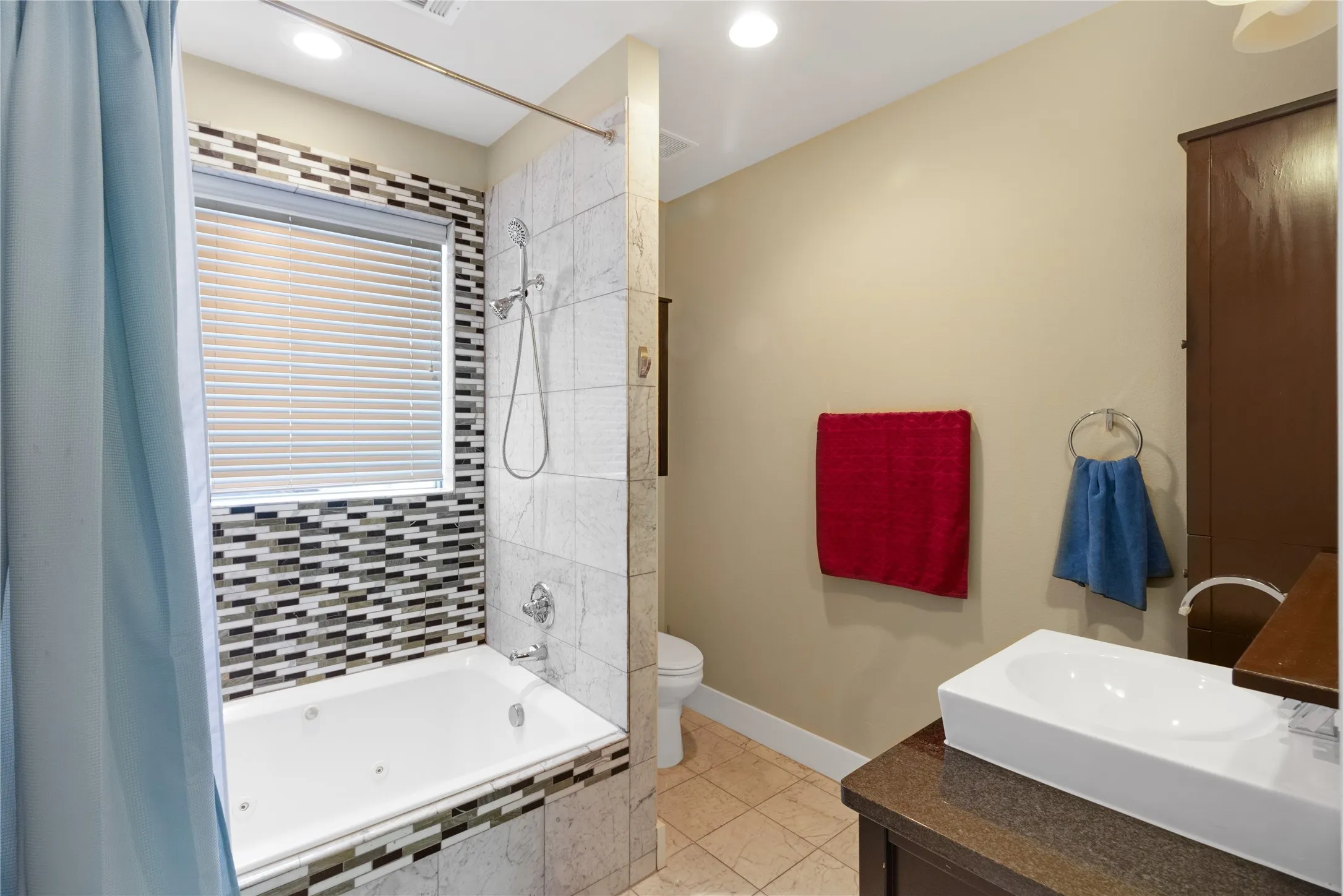 Bathroom featuring a built-in tub with mosaic tile surround, an overhead shower, a vessel sink on a dark vanity, and neutral wall tones