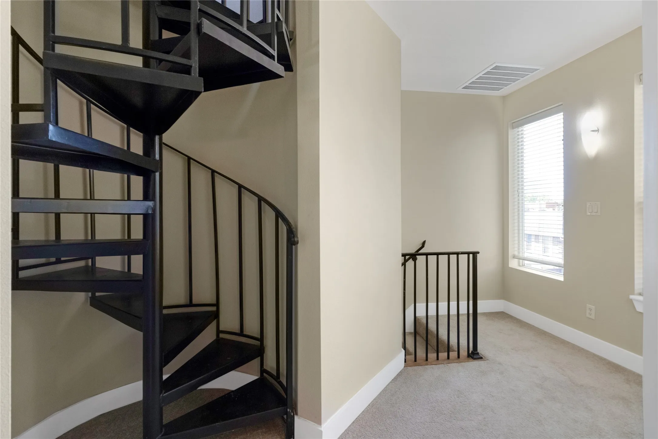 Black metal spiral staircase featuring a continuous handrail, leading to a carpeted landing with a window