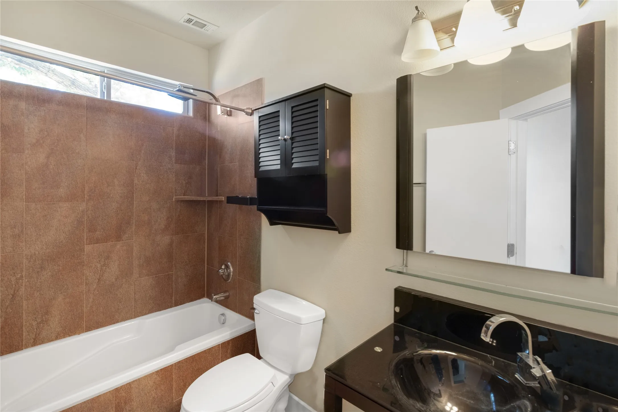 Bathroom featuring a built-in tub with tile surround, a dark wood-finish vanity with an integrated basin, an accent mirror with integrated lighting, and a wall-mounted storage cabinet