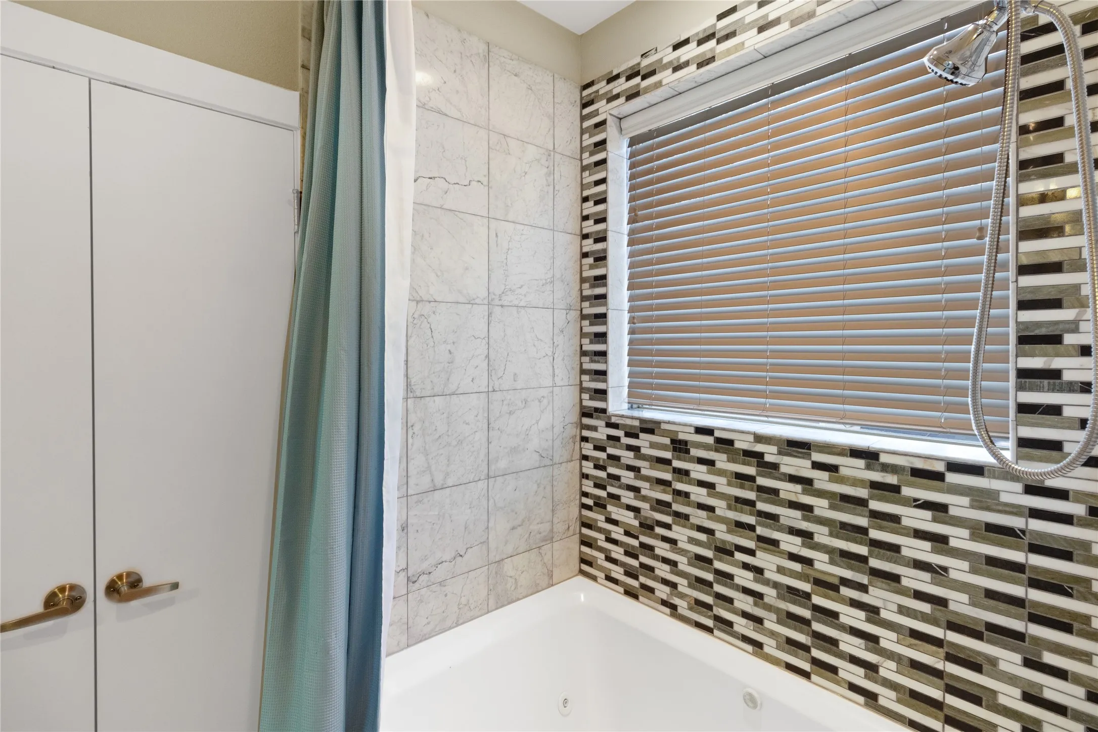 Bathtub with a mosaic tile surround featuring a window with horizontal blinds, complemented by a handheld shower fixture