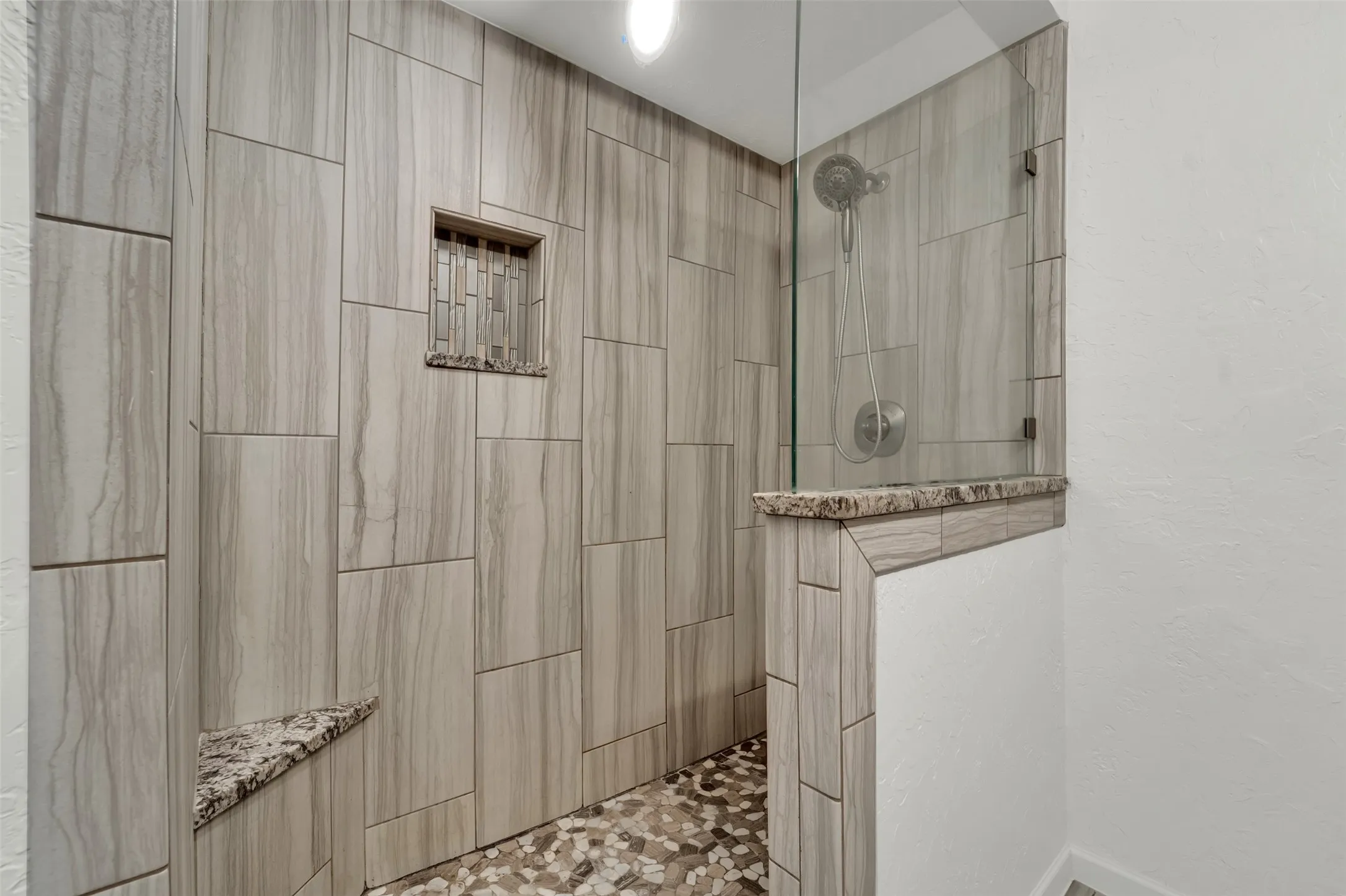 Shower featuring large format wood-grain tile, a built-in niche with mosaic tile, a granite-topped half wall, a frameless glass enclosure, and a tiled shower bench