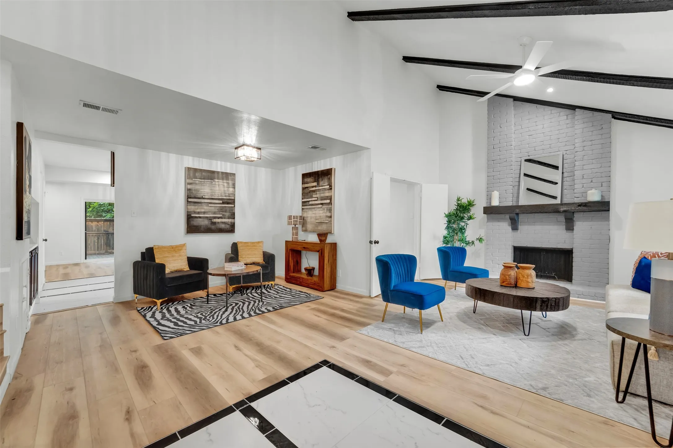 Vaulted ceiling with exposed dark beams, light wood-finish flooring, painted brick fireplace with dark wood mantel, white walls, and a ceiling fan