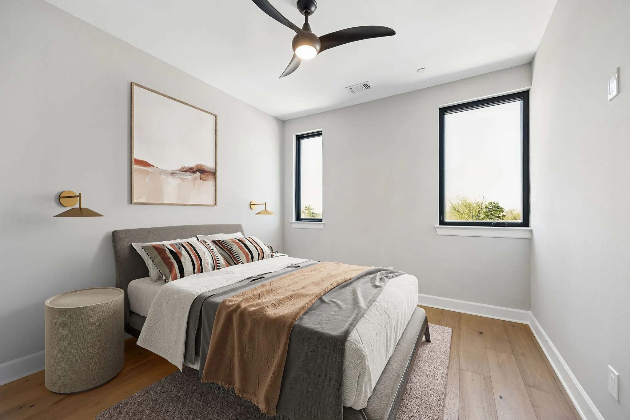 Hardwood flooring, two rectangular windows with black frames, a ceiling fan with integrated lighting, and two wall-mounted sconces