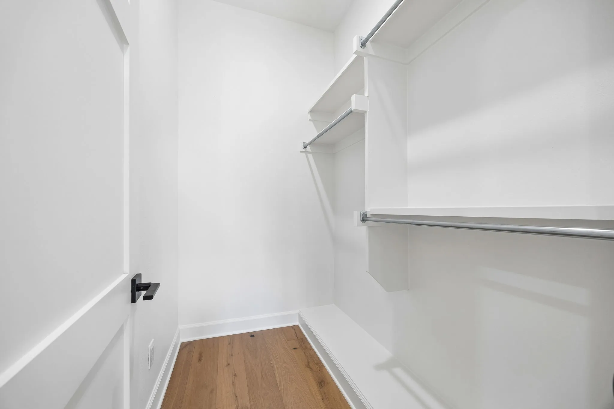 Walk-in closet featuring white walls, wood-finish flooring, white baseboards, and built-in white shelving with metal hanging rods