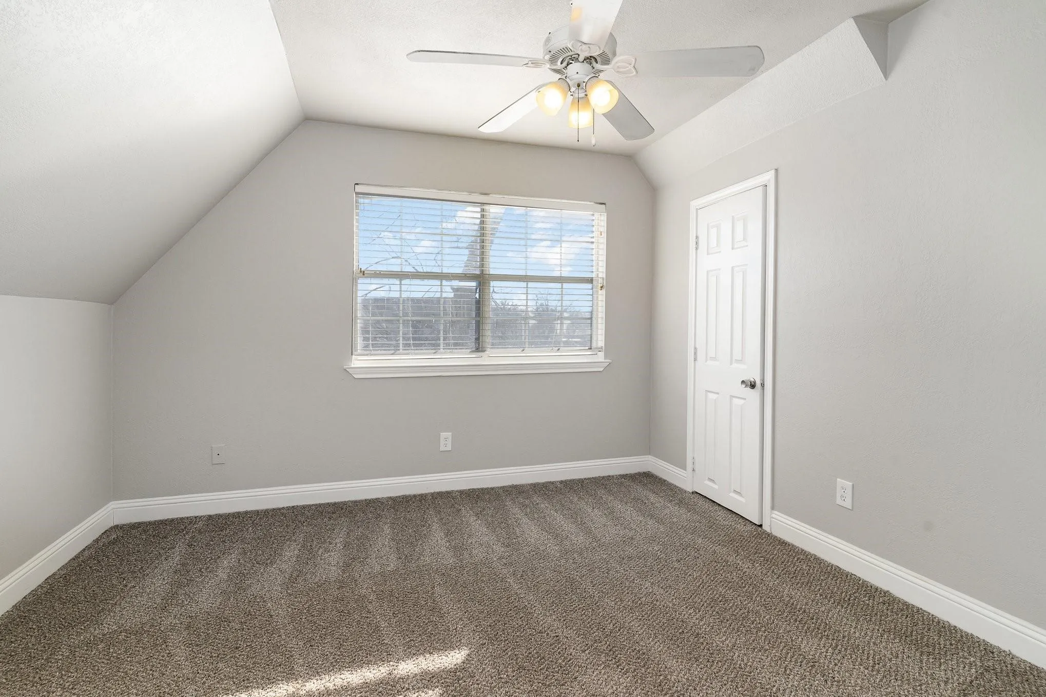 Carpeted room featuring a window with blinds, a white ceiling fan with integrated lighting, a paneled door, and angled ceilings